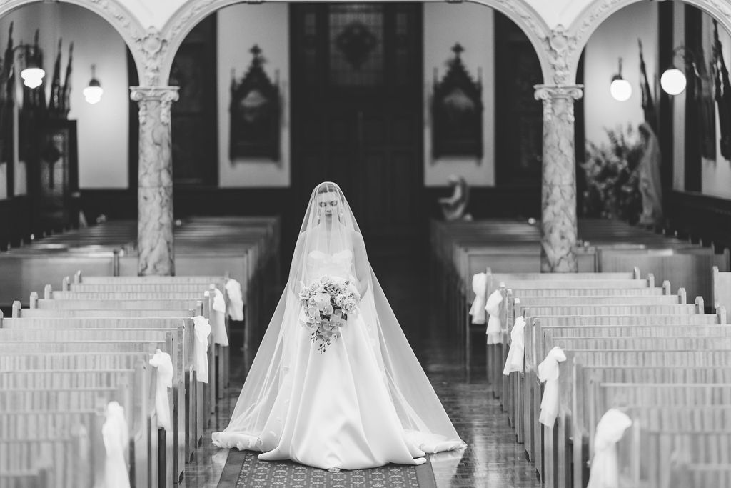 Black and white photo of a bride in a flowing gown walking down a church aisle holding a bouquet at All Hallows' School Queensland