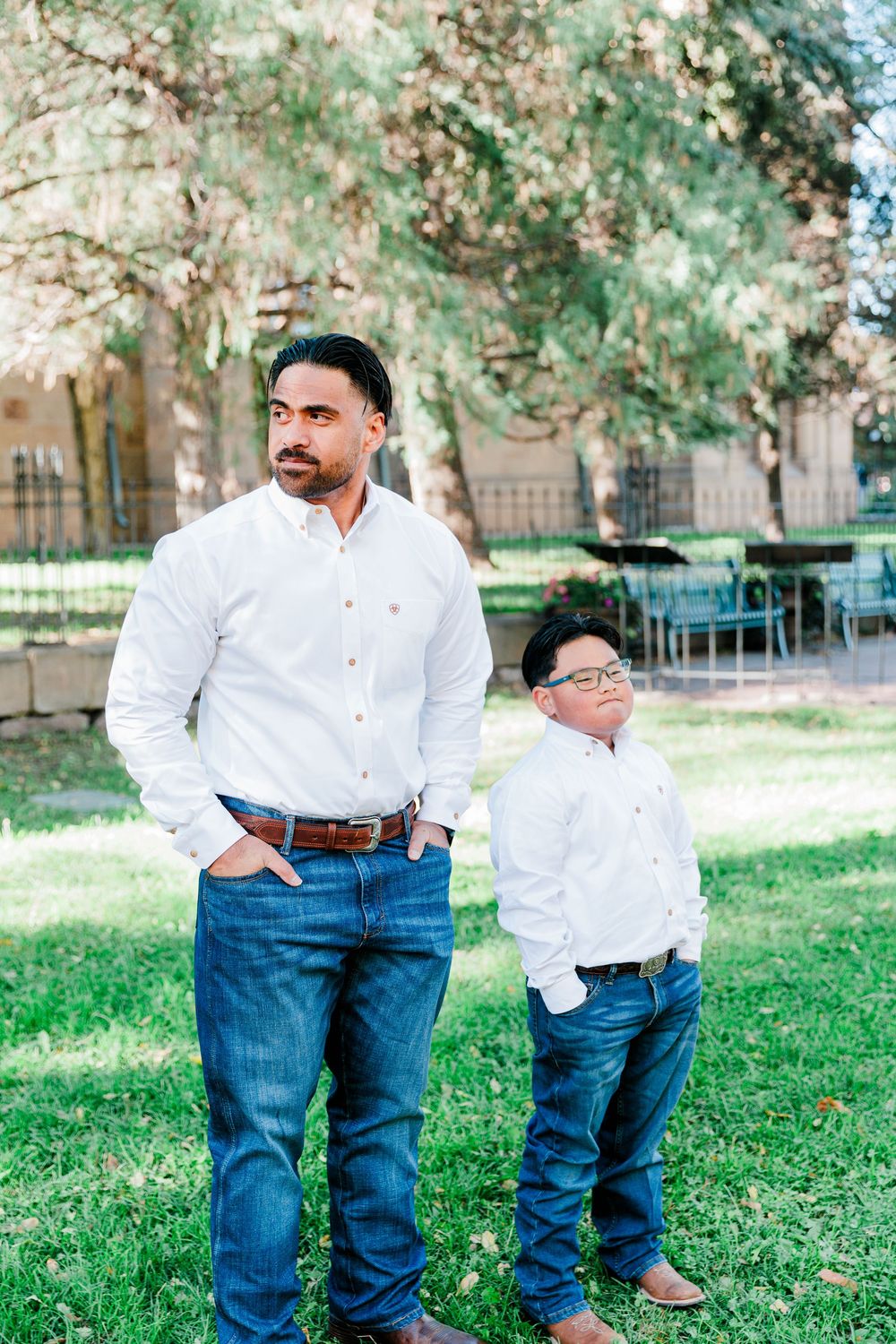 A man and a young boy in matching white button-up shirts and blue jeans stand on a grassy lawn under trees.