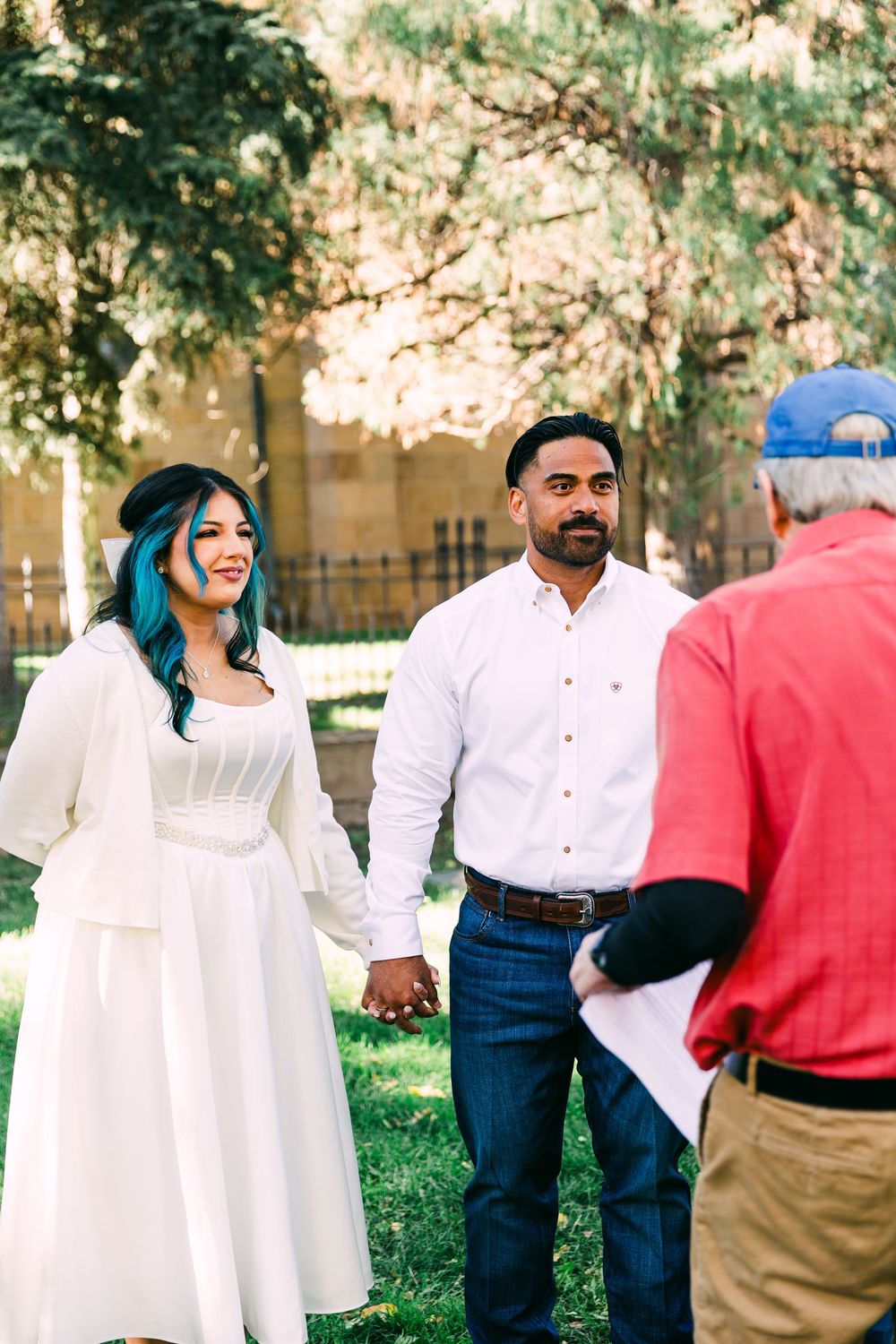 Bride with teal-blue hair in a white dress holds hands with a groom in a white shirt during an outdoor wedding ceremony in a sunlit park.