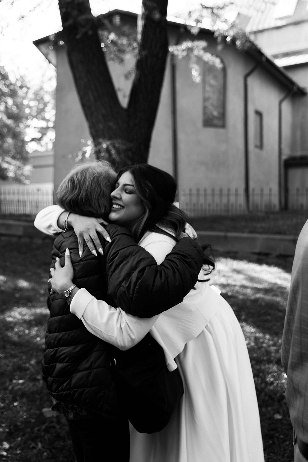 Black and white photo of a smiling bride in a white dress warmly hugging a guest in a puffer jacket outside a historic building.