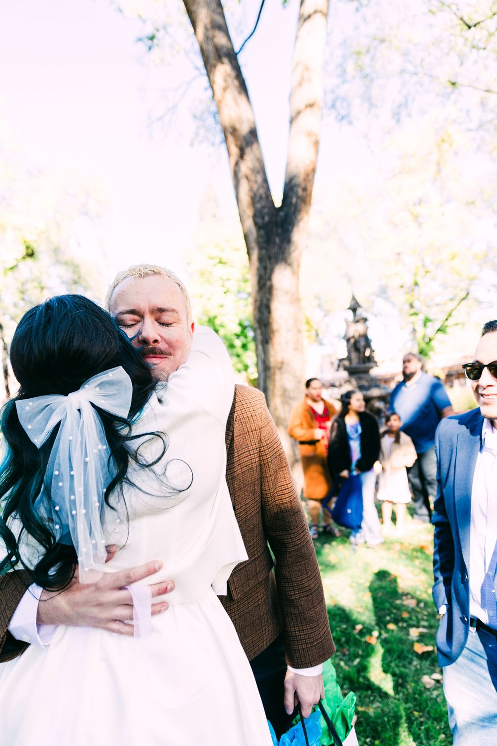 Bride in a white dress with a tulle bow embraces an emotional man in a tweed jacket during an outdoor wedding as guests gather under trees.
