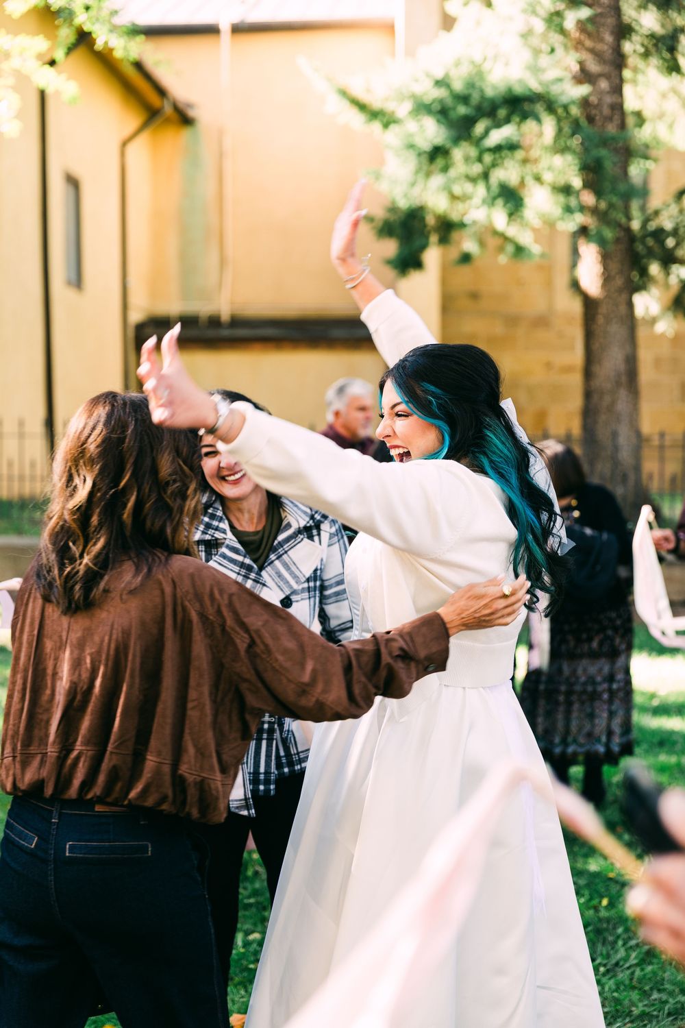 A bride with teal-streaked hair in a white gown raises her arms and laughs while dancing with guests at an outdoor wedding.