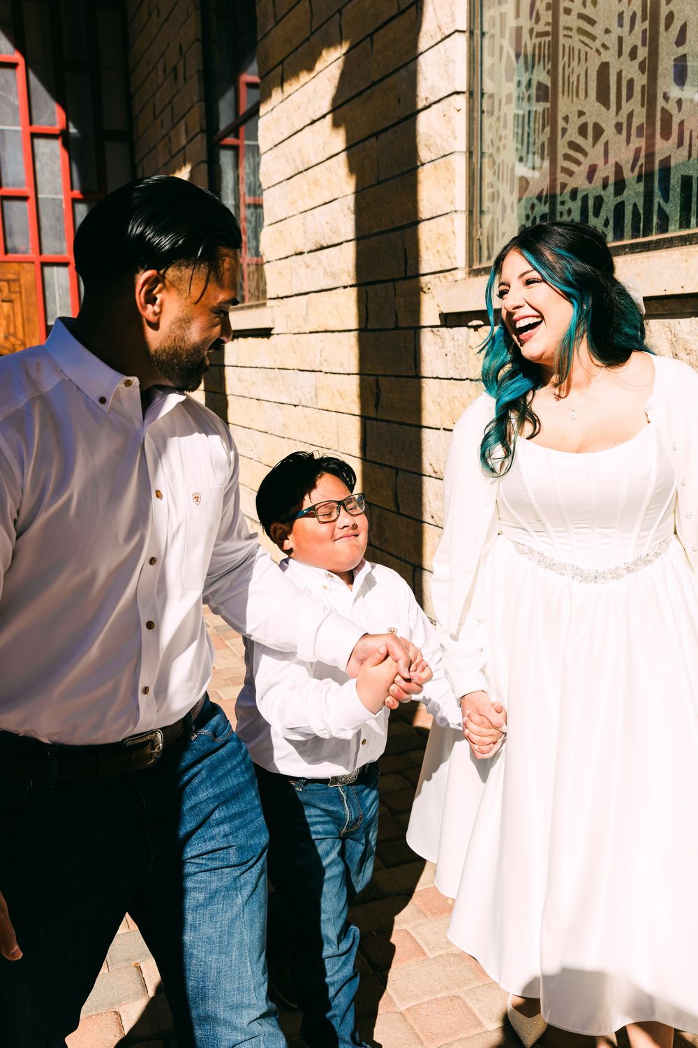 A laughing bride with teal-dyed hair in a white dress walks hand in hand with a smiling boy and a man in jeans beside a brick building.