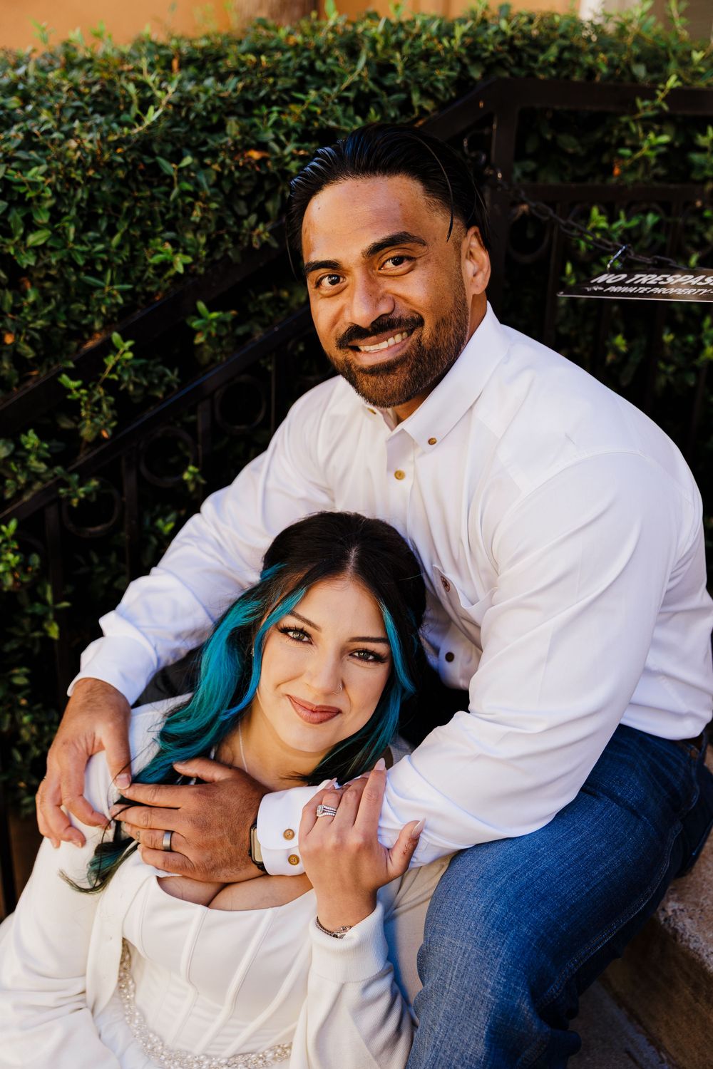 A smiling man in a white shirt embraces a woman with teal-dyed hair in a white dress while posing on outdoor steps by a hedge.