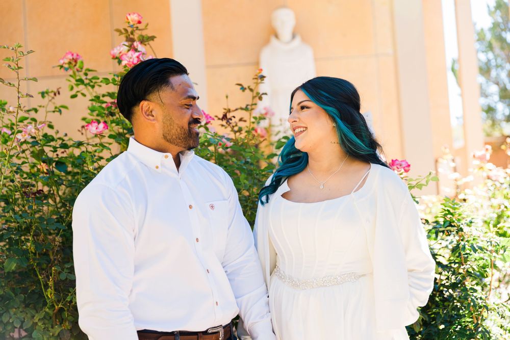 Smiling couple in white outfits poses close together among pink rose bushes with a classical statue and warm stone architecture behind them.