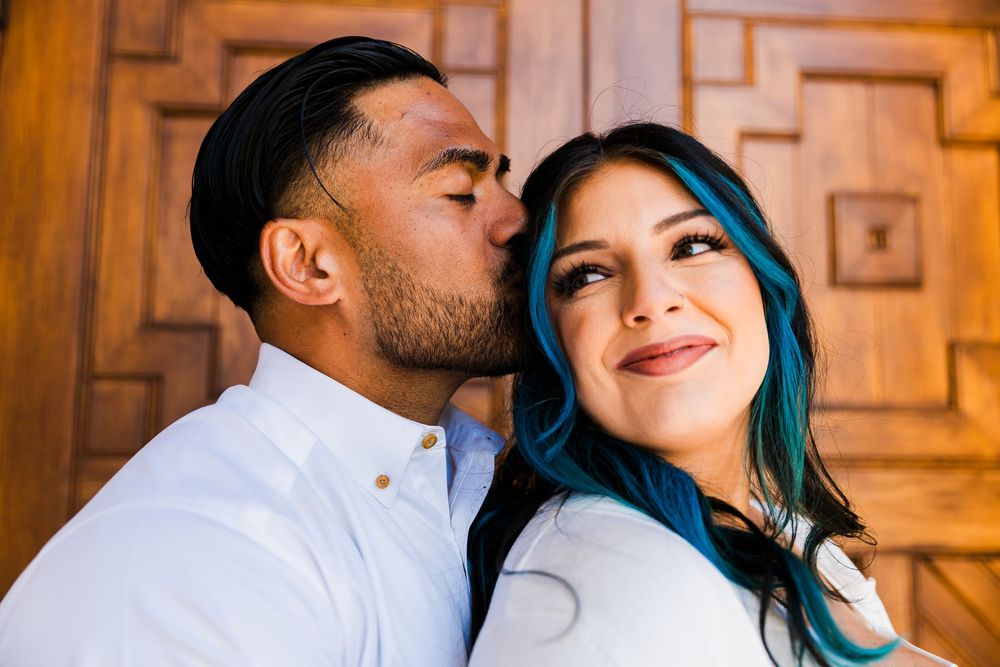Man in a white button-up shirt kisses the cheek of a smiling woman with blue-streaked dark hair in front of a carved wooden door.