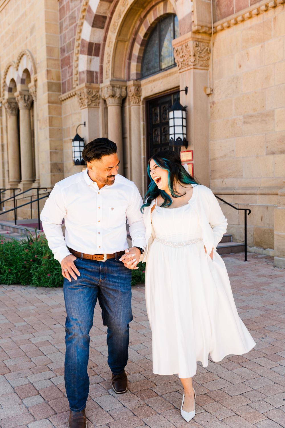 Smiling couple in white outfits and jeans walk hand in hand across a brick plaza in front of a historic stone building with arched doorways.
