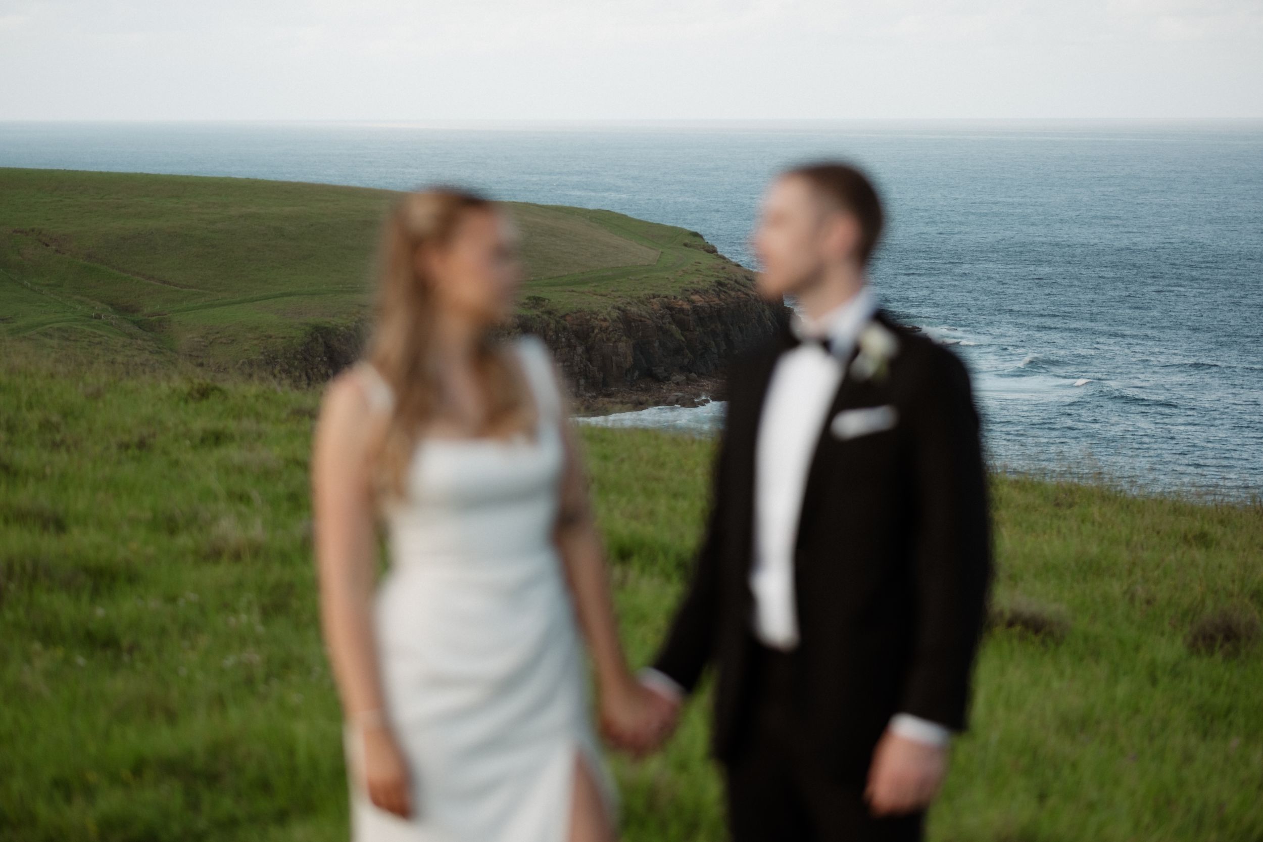 A bride and groom hold hands on a lush green coastal cliff, with ocean waves and rugged headlands in the background.