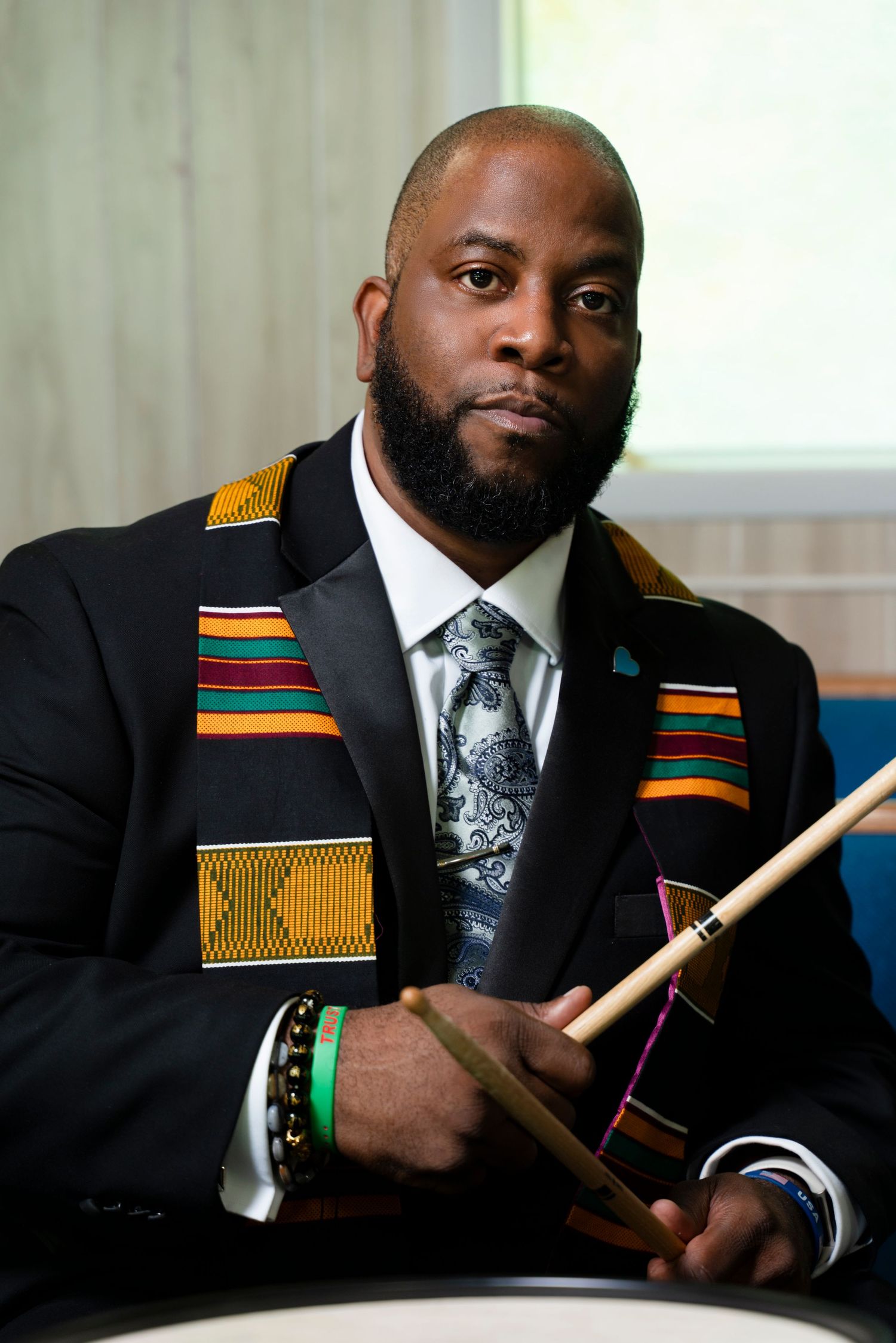 Man in black suit with kente cloth stole holding a Holy Bible, standing before a brick wall.