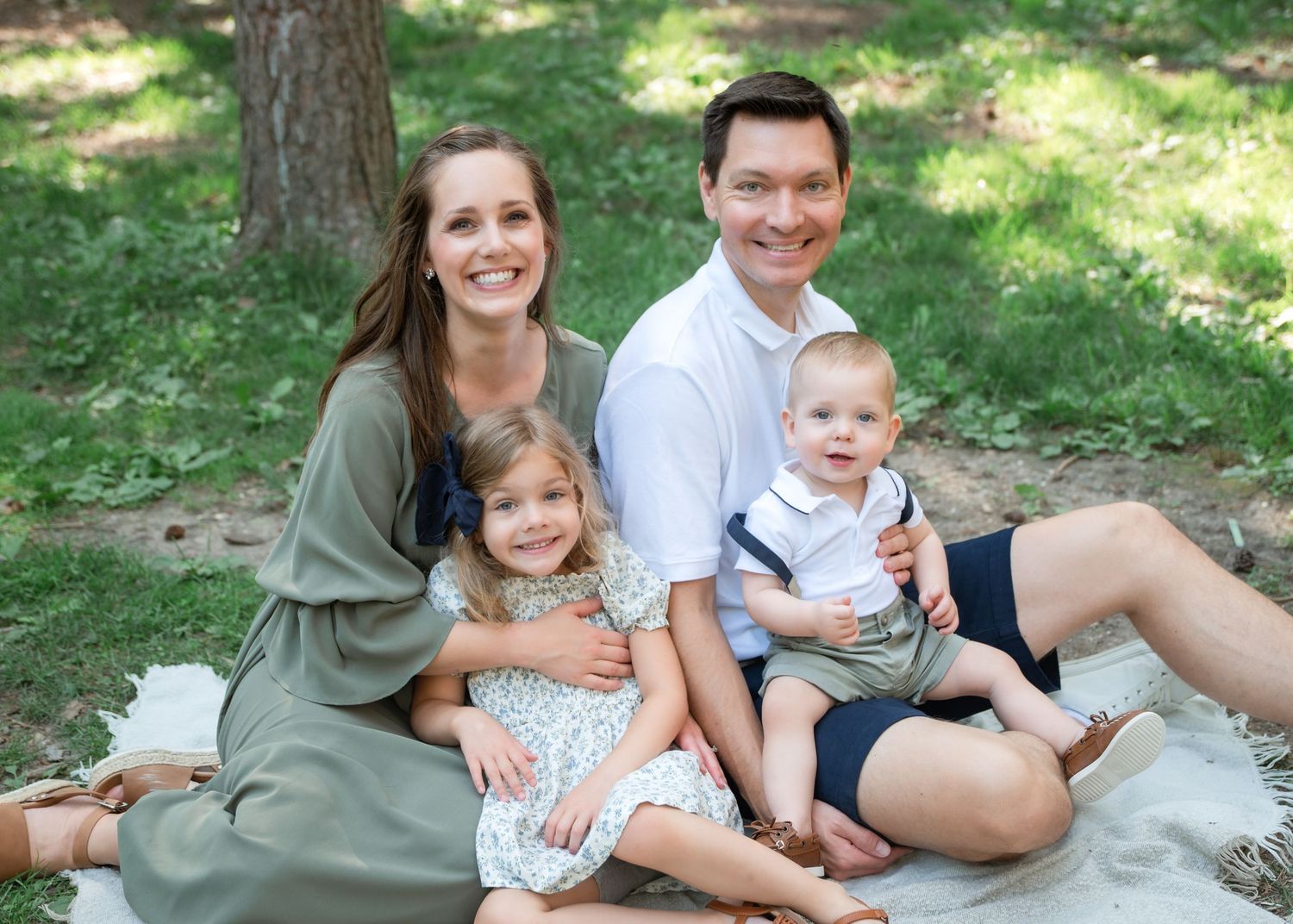 Happy family of four smiling outdoors on a blanket in a lush green park setting.