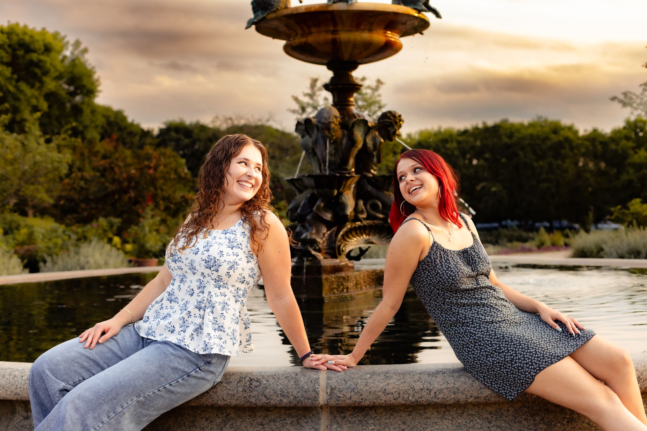 Two women smile and pose by an ornate bronze fountain at sunset in a lush park setting.