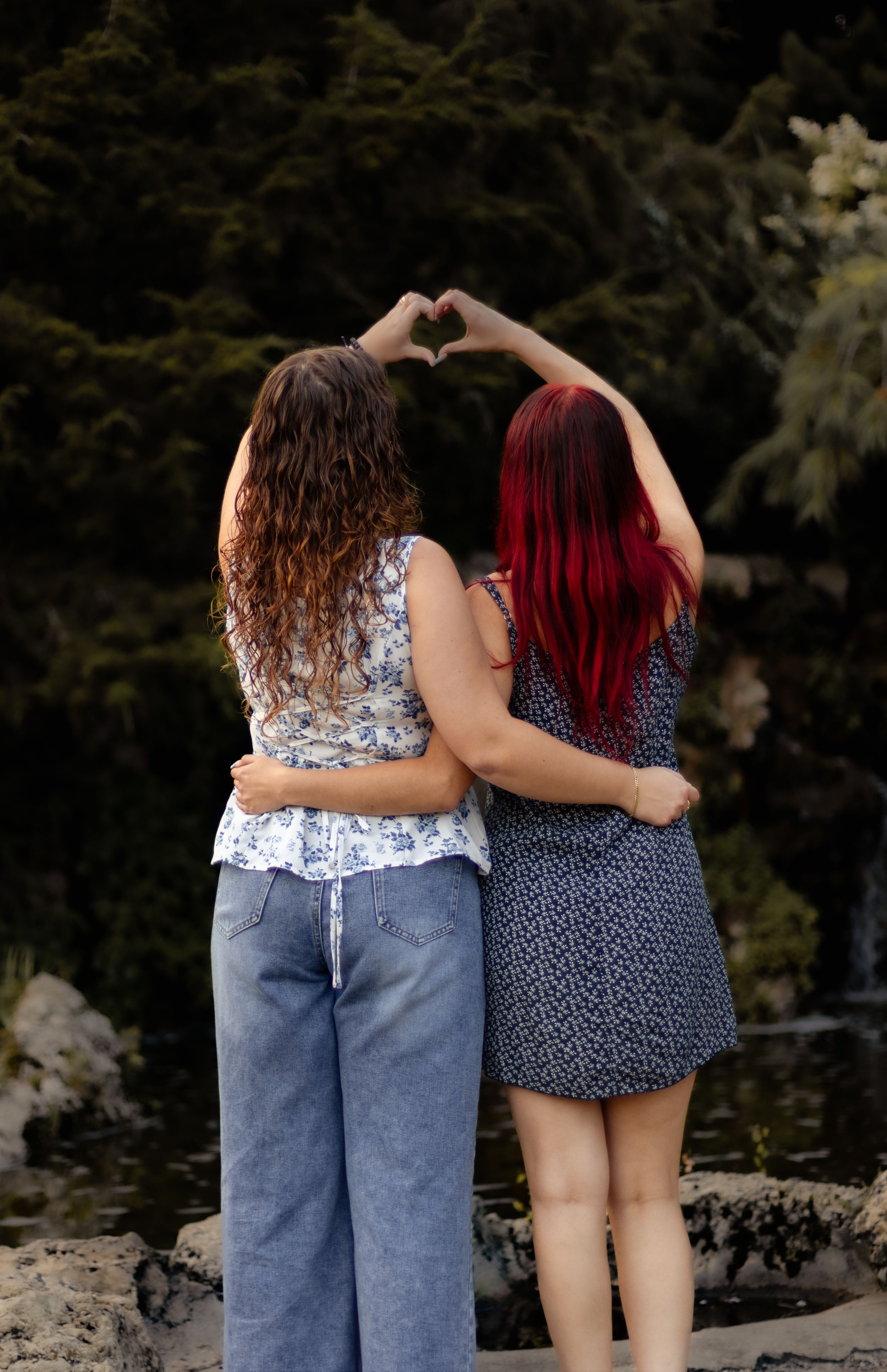Two women with curly and red hair make a heart shape with their hands while embracing outdoors near a pond.