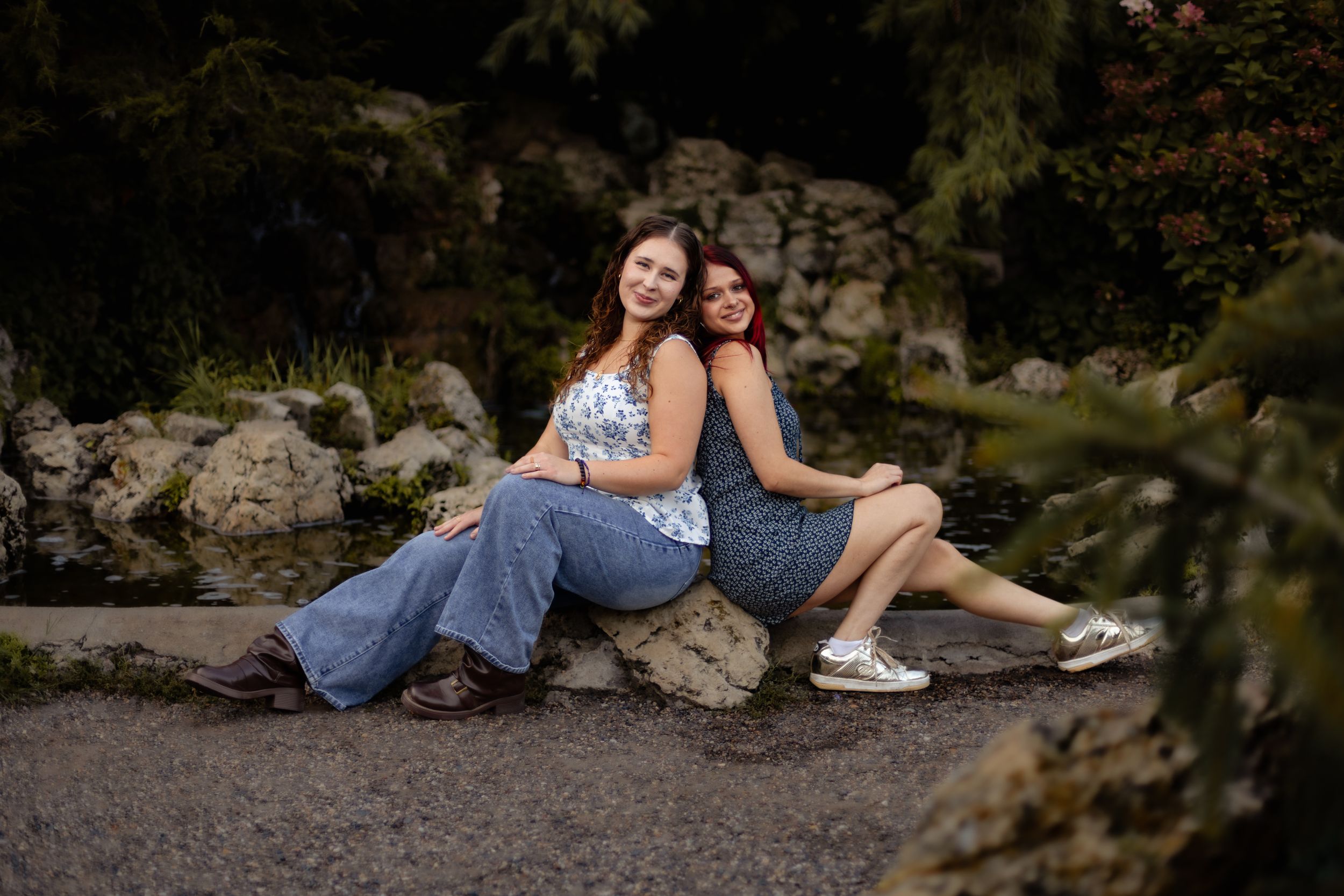 Two young women sitting back-to-back on a rock in a lush garden, wearing casual blue outfits, smiling warmly.