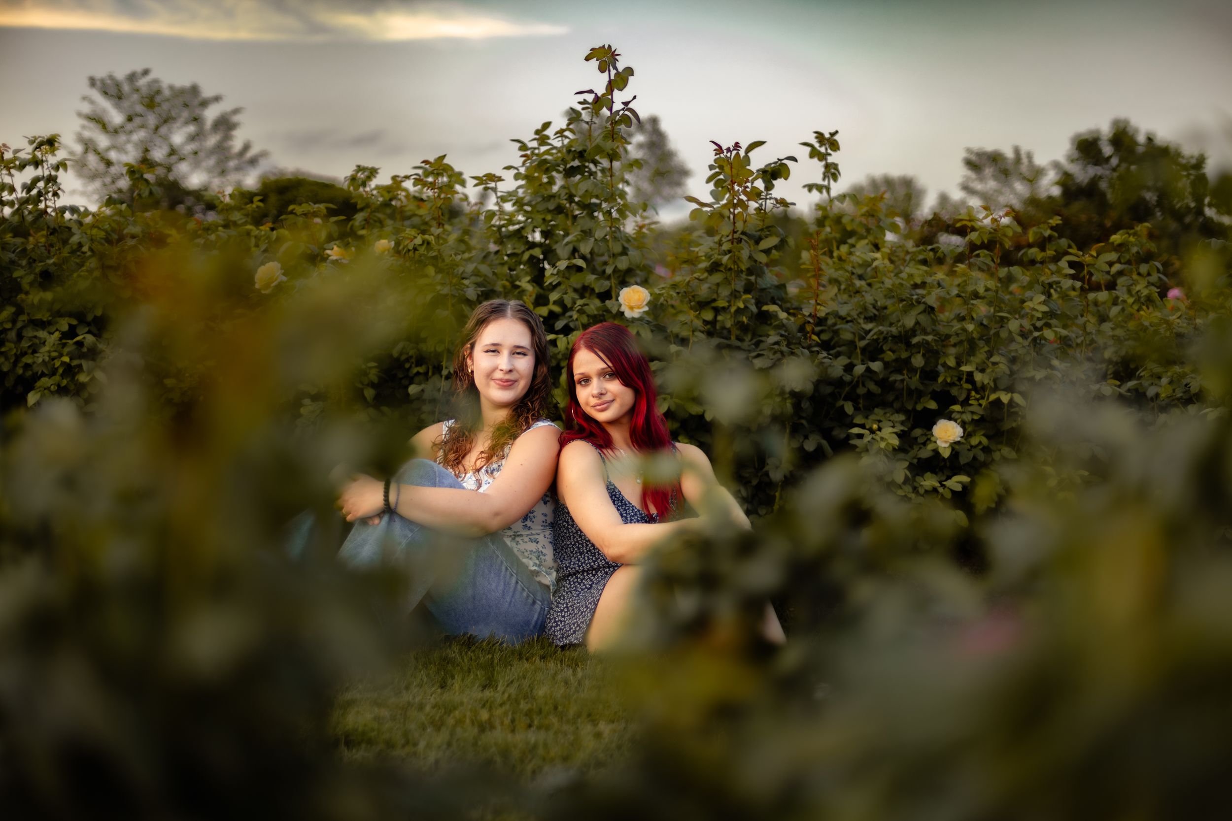 Two young women sitting together among lush green rose bushes in a garden at golden hour.