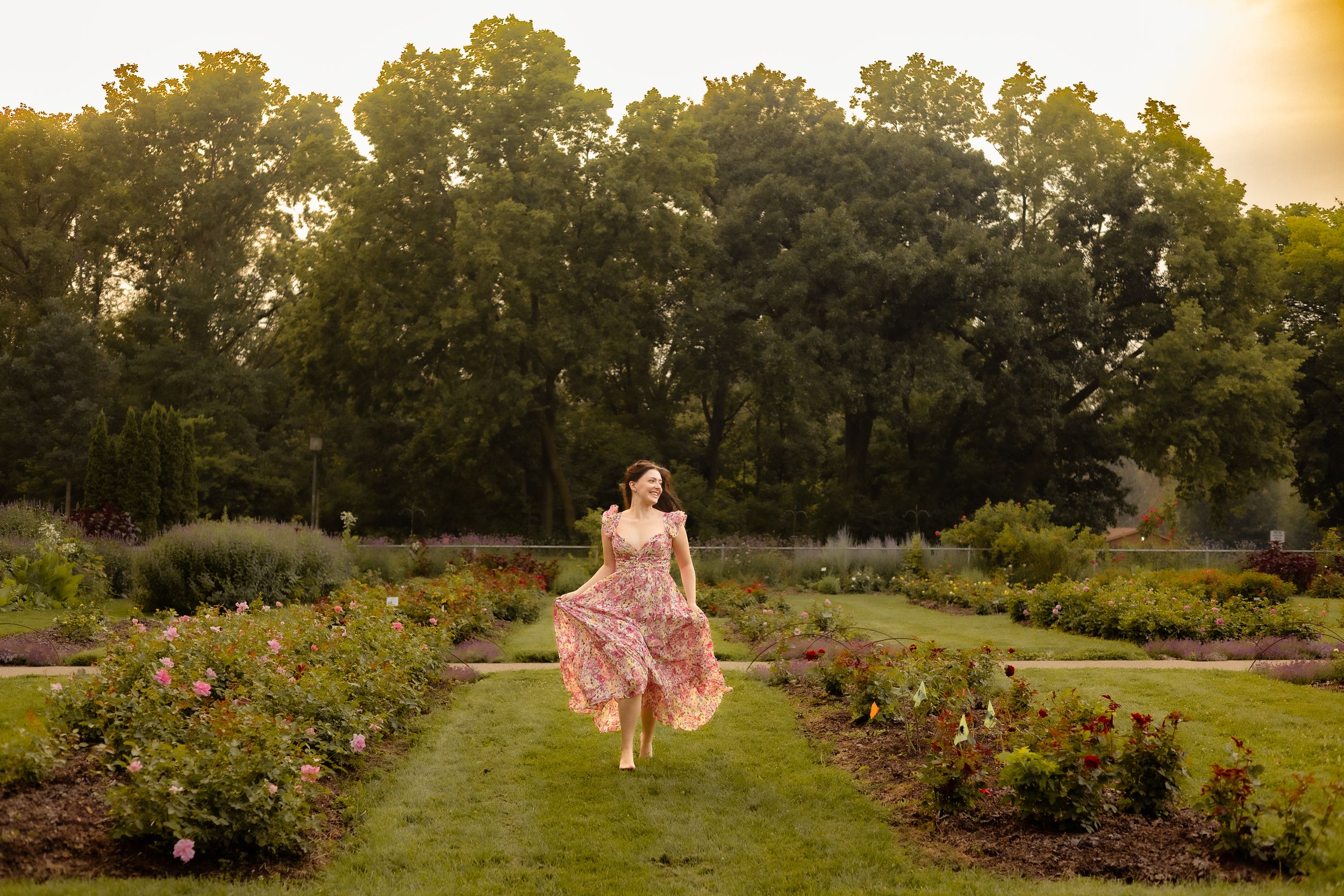 Woman in floral dress twirling in a lush garden at golden hour with tall green trees in the background.