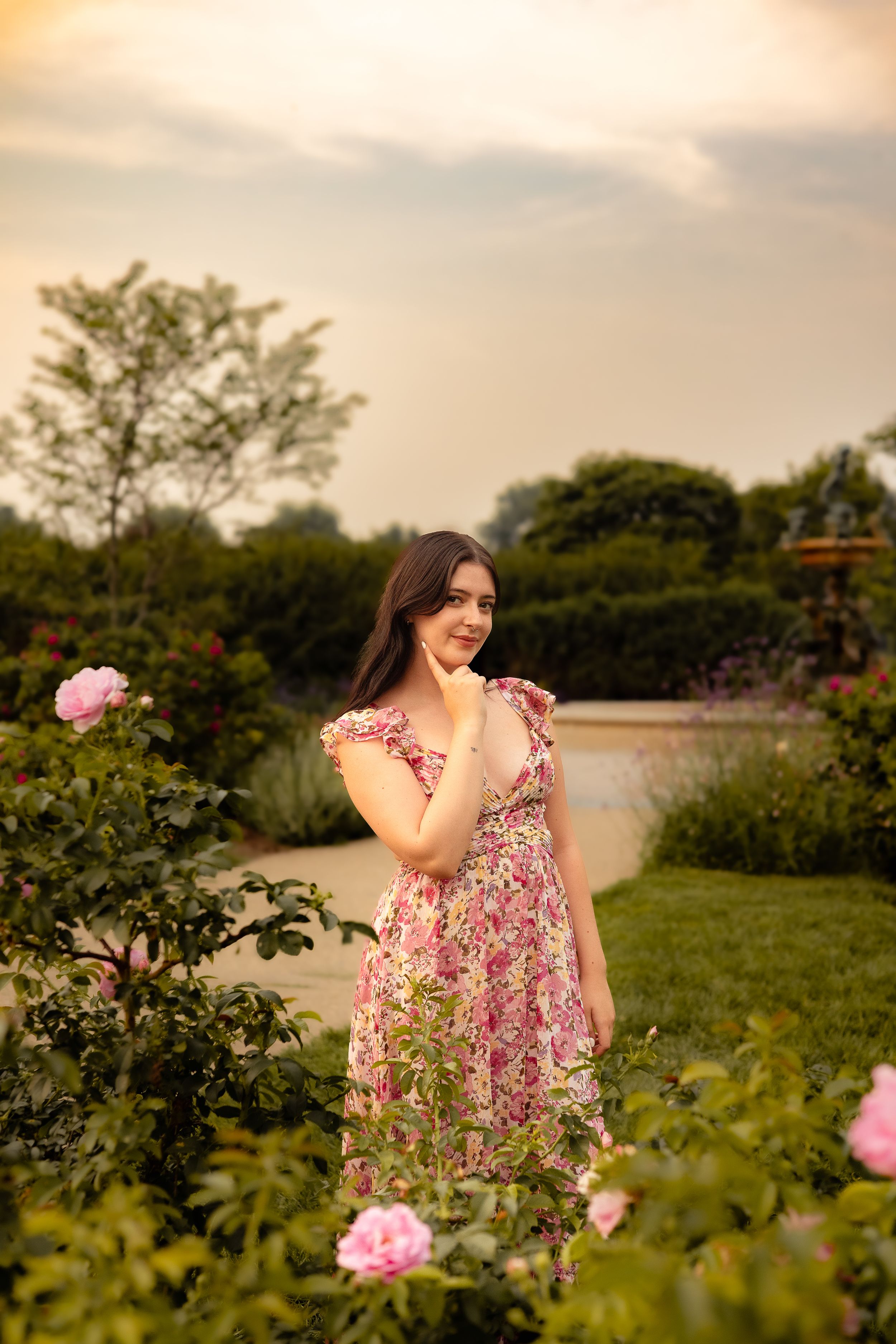 Young woman in floral dress smiling among pink roses in a lush garden at sunset.
