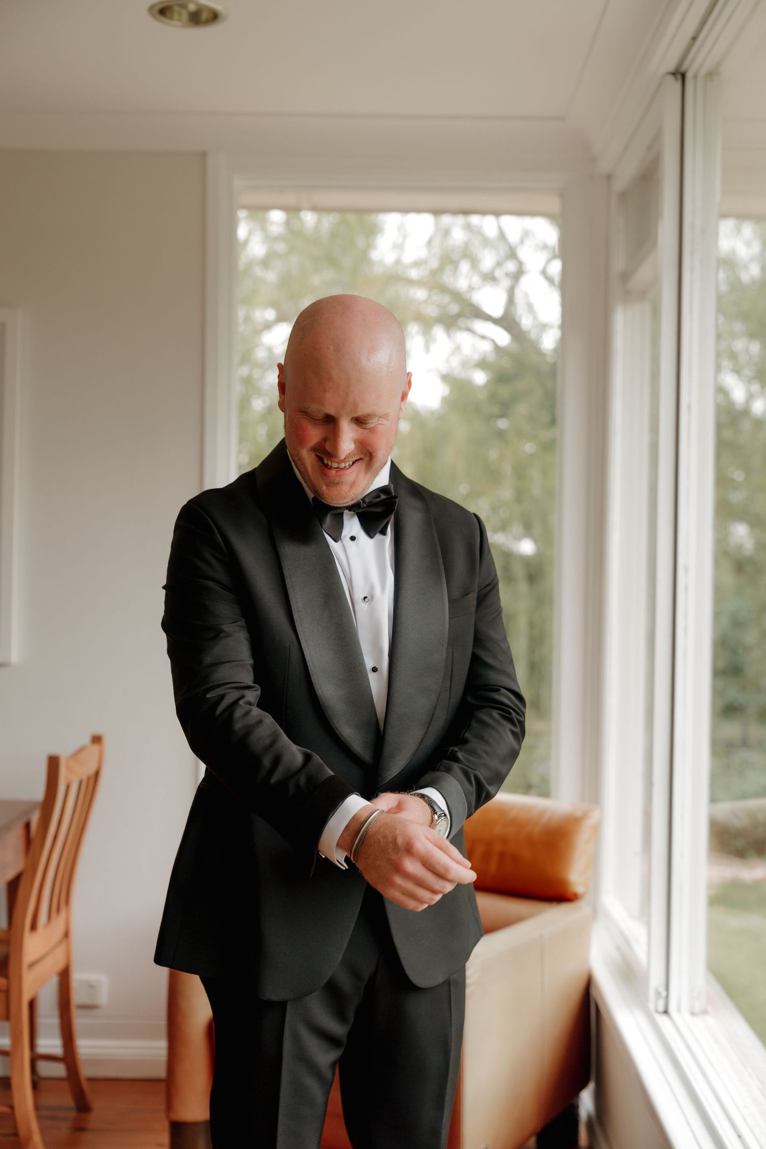 Smiling bald groom in black tuxedo and bow tie adjusting cufflinks by window before wedding ceremony. The Vinegrove Mudgee. Mudgee Wedding