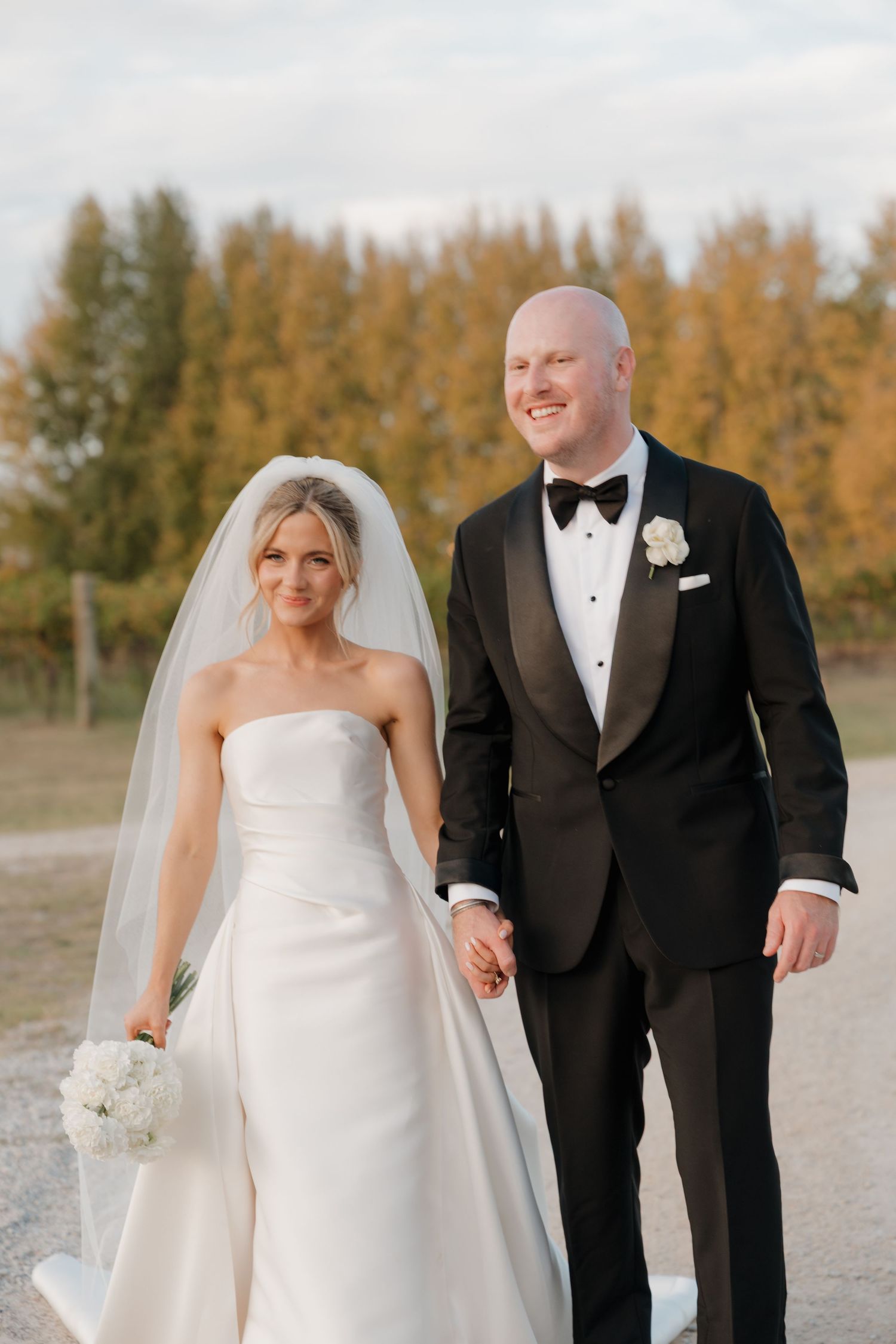 Bride in strapless white gown and veil holds bouquet while walking hand-in-hand with groom in black tuxedo outdoors.