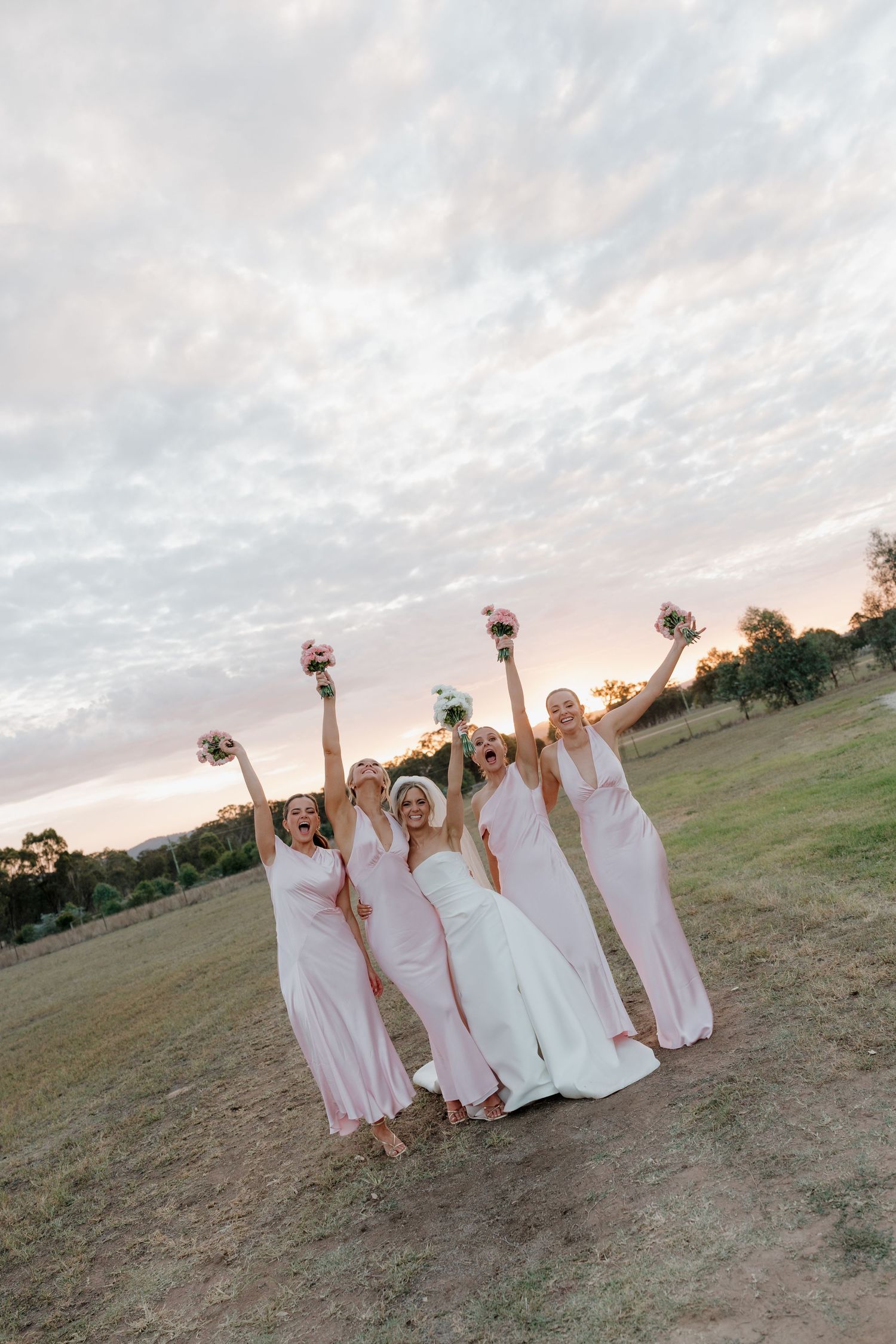 Bride and bridesmaids in pink dresses celebrating outdoors at sunset, raising bouquets and cheering on a green hillside.