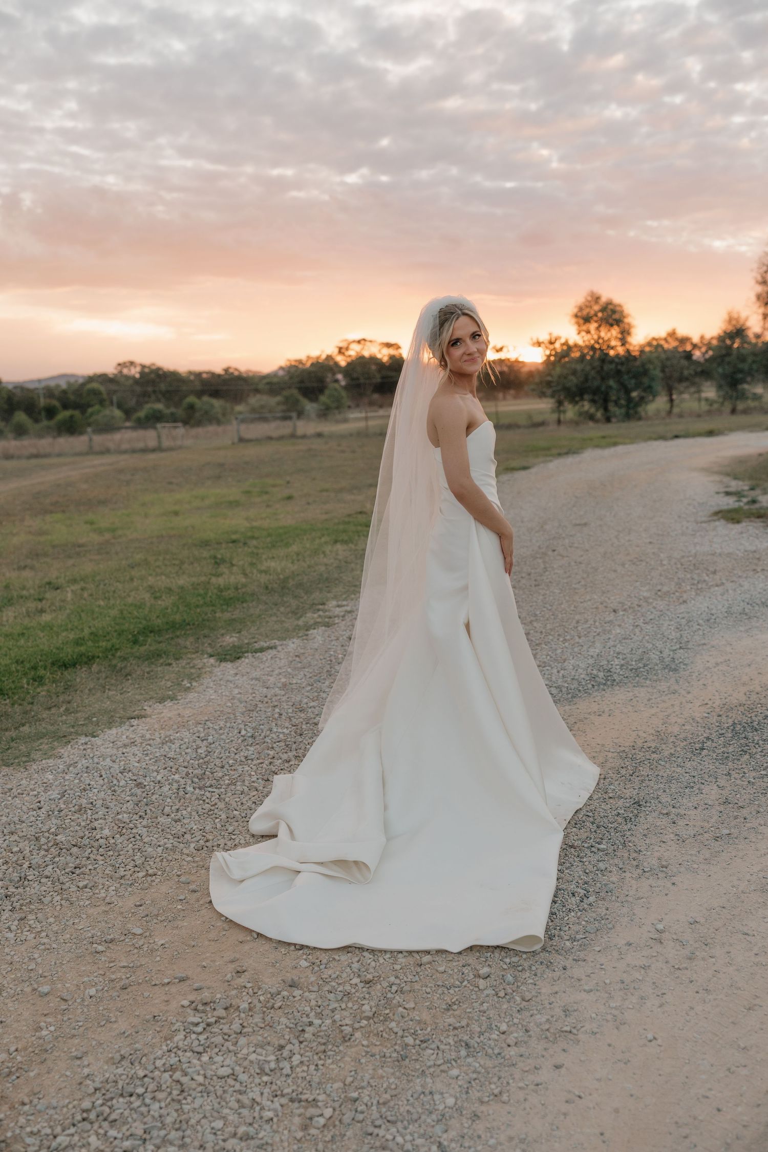 Bride in white strapless gown with long veil stands on gravel path at golden sunset. The Vinegrove Mudgee. Mudgee Wedding Photographer