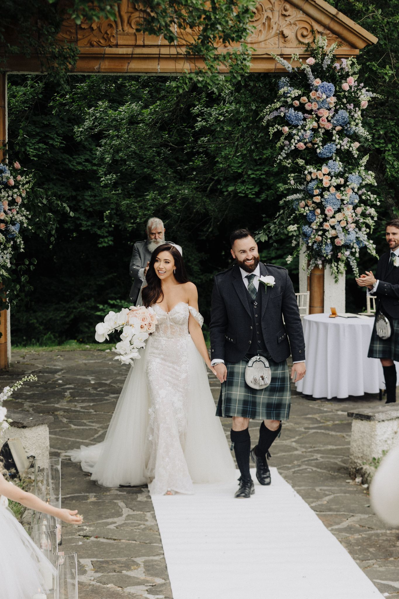 Newlyweds walking down the aisle at an outdoor ceremony decorated with blue and pink flowers.