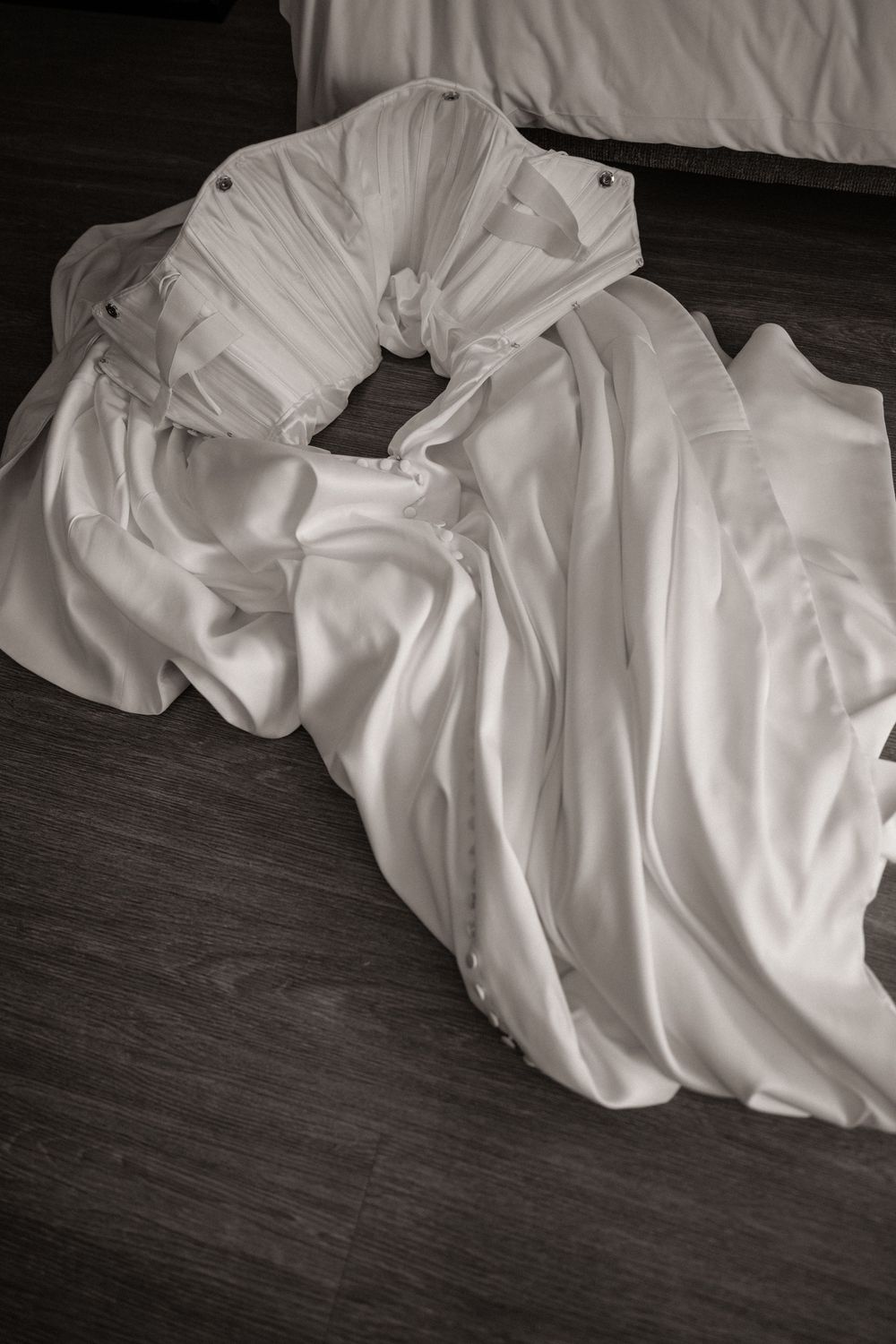 Black and white overhead view of white satin wedding dress laid out on wooden floor.