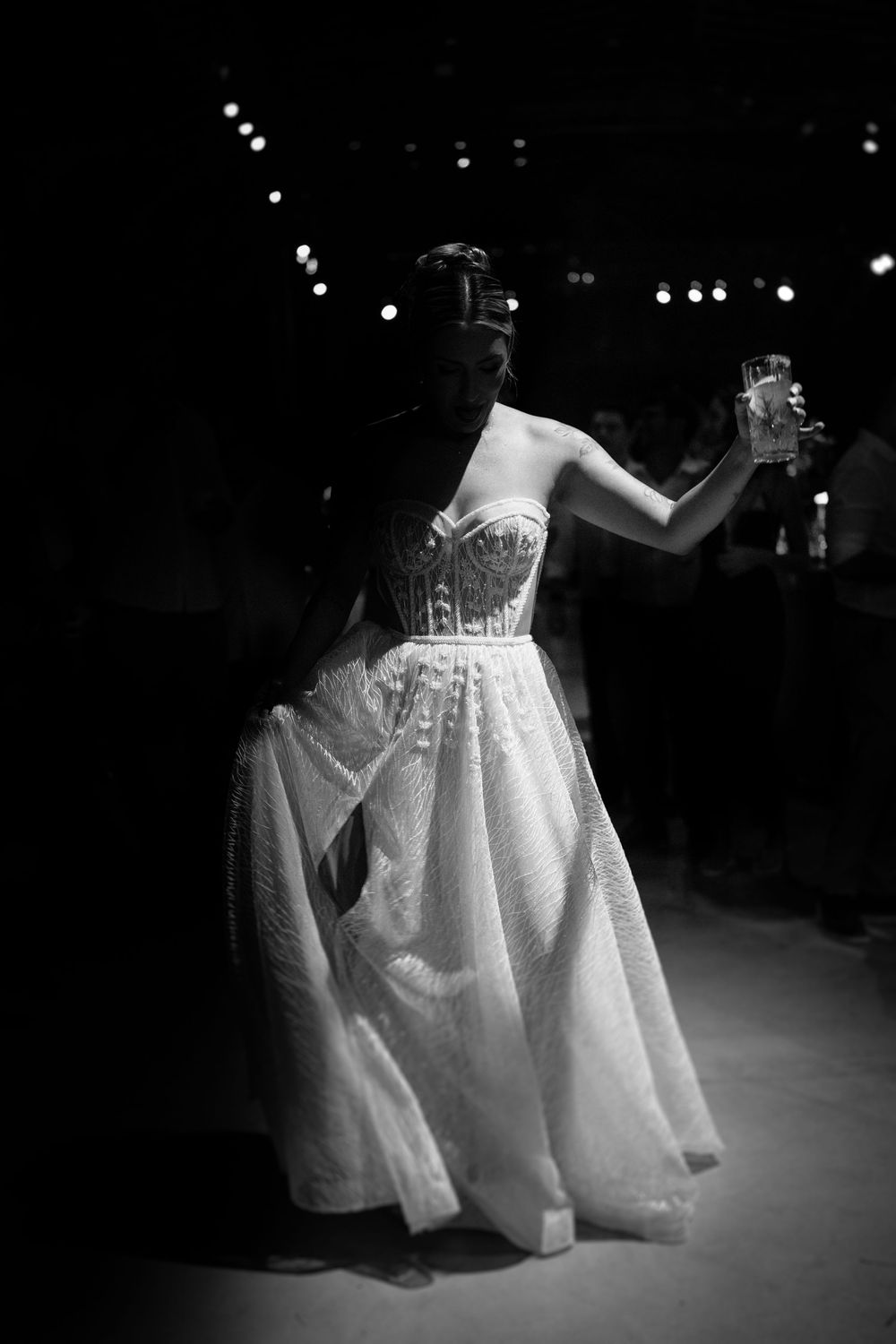 Black and white photo of bride in strapless corset ball gown raising glass at dark reception.