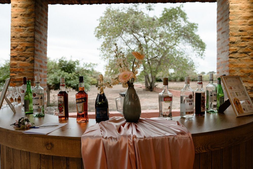 Elegant outdoor wedding bar setup with bottles and floral arrangement on draped table.