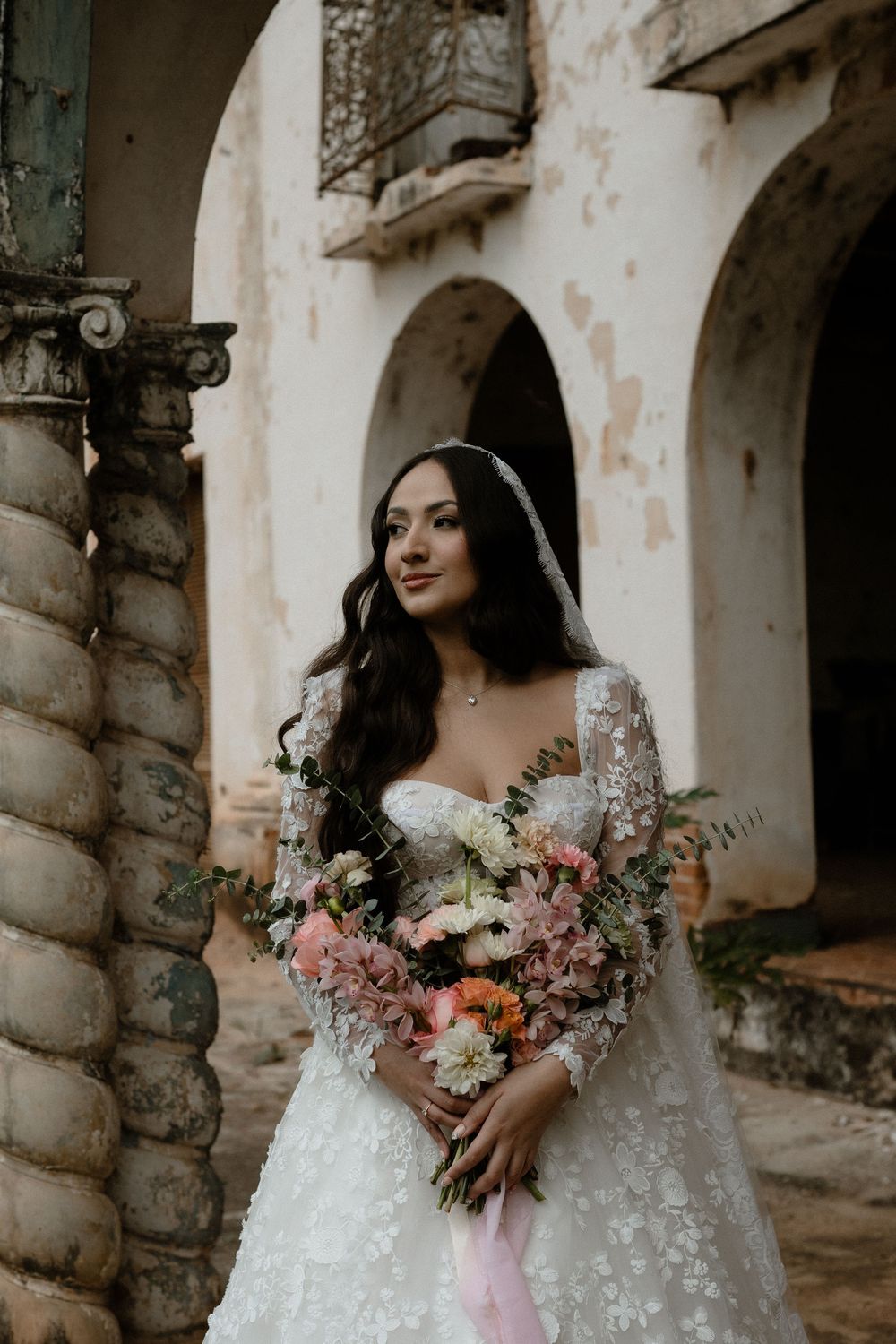 Bride in floral lace gown with veil holds pink bouquet before rustic colonial architecture.
