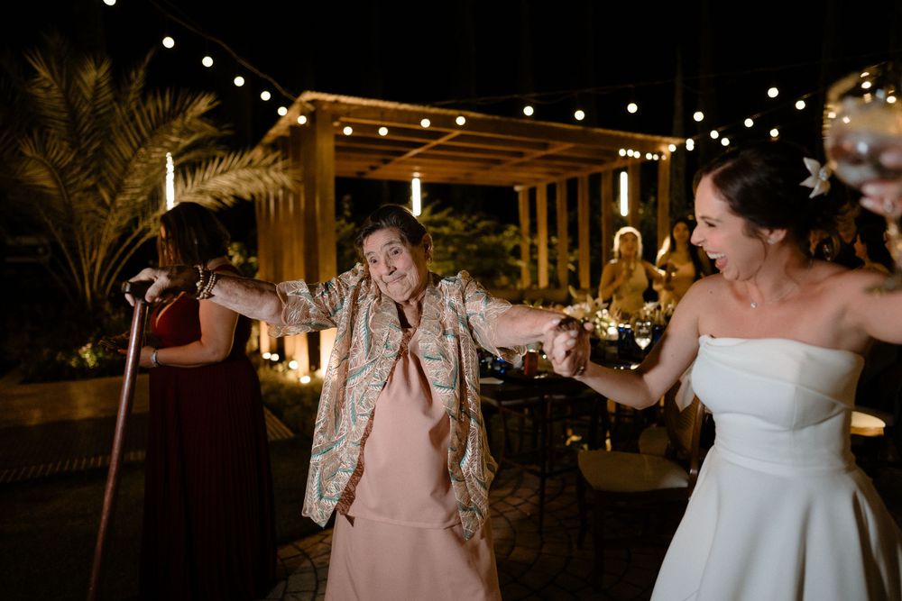 Joyful bride dancing with elderly guest under string lights at outdoor wedding reception.