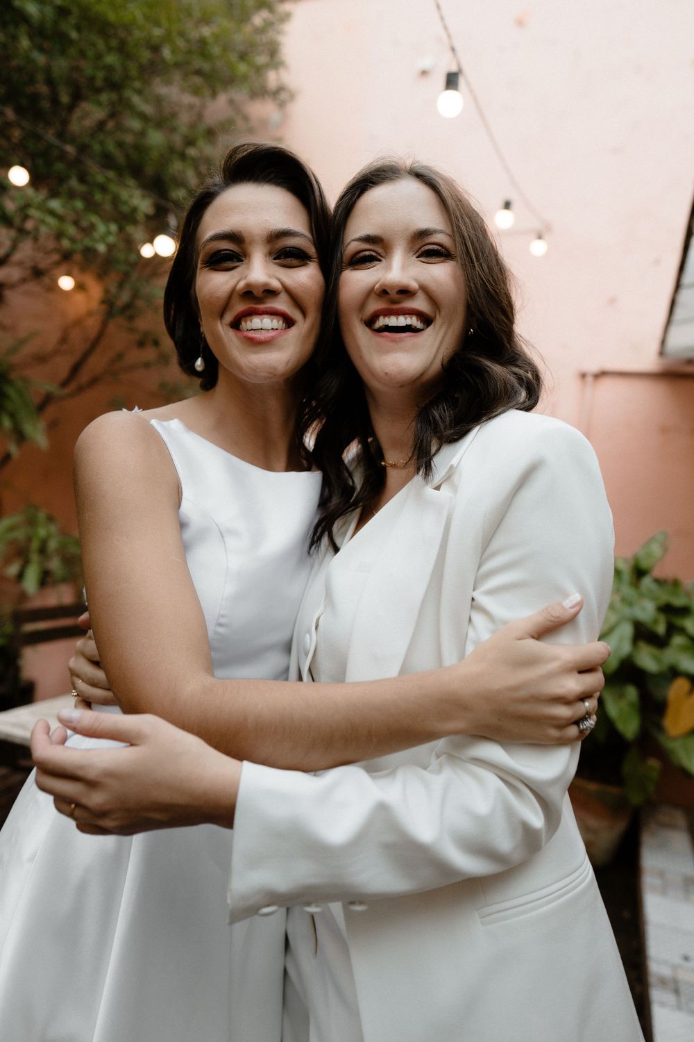 Two brides in white embrace and smile joyfully in a courtyard decorated with string lights at their wedding.