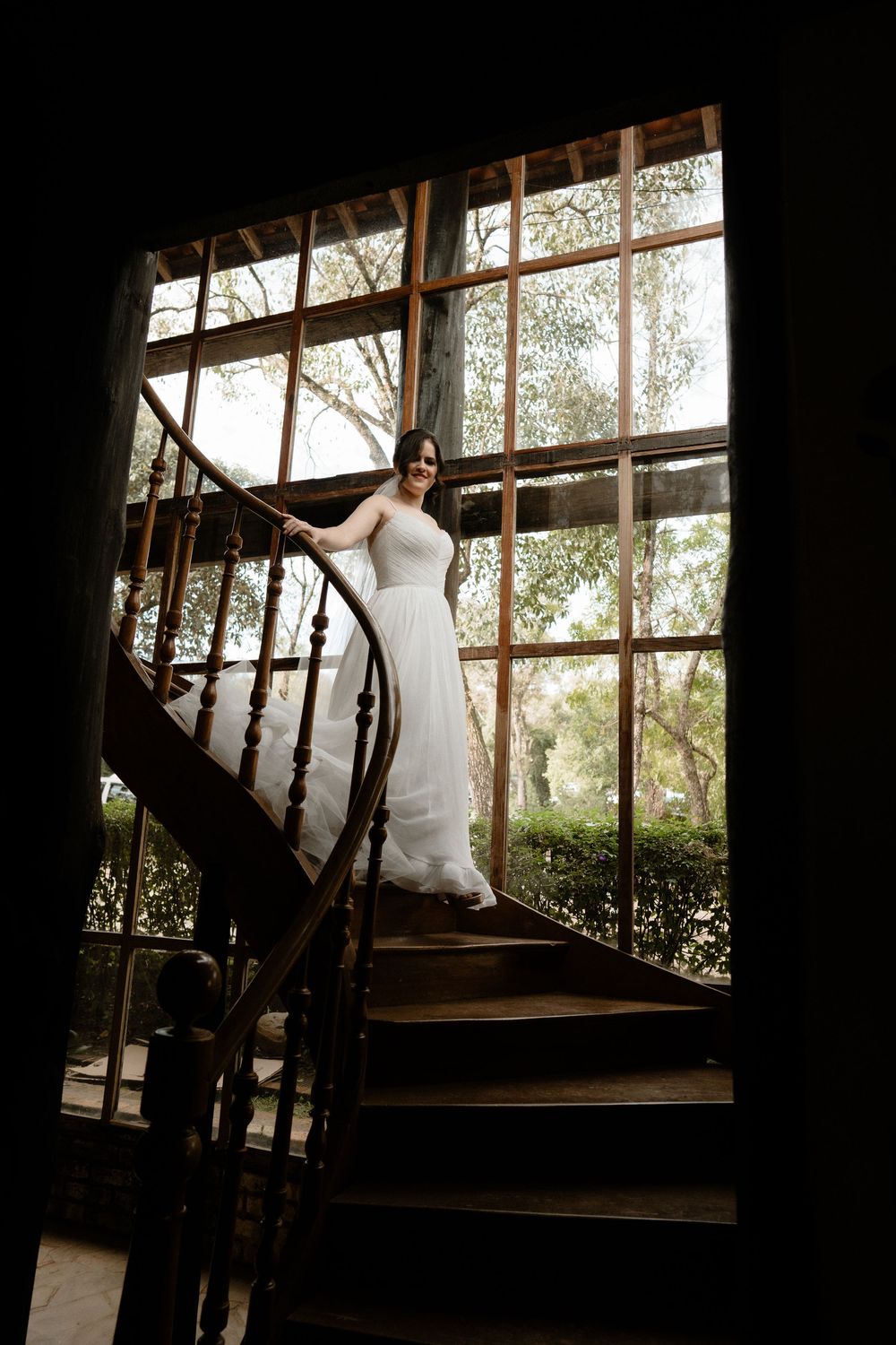 Bride in white gown standing on spiral staircase by large window in sunlight.