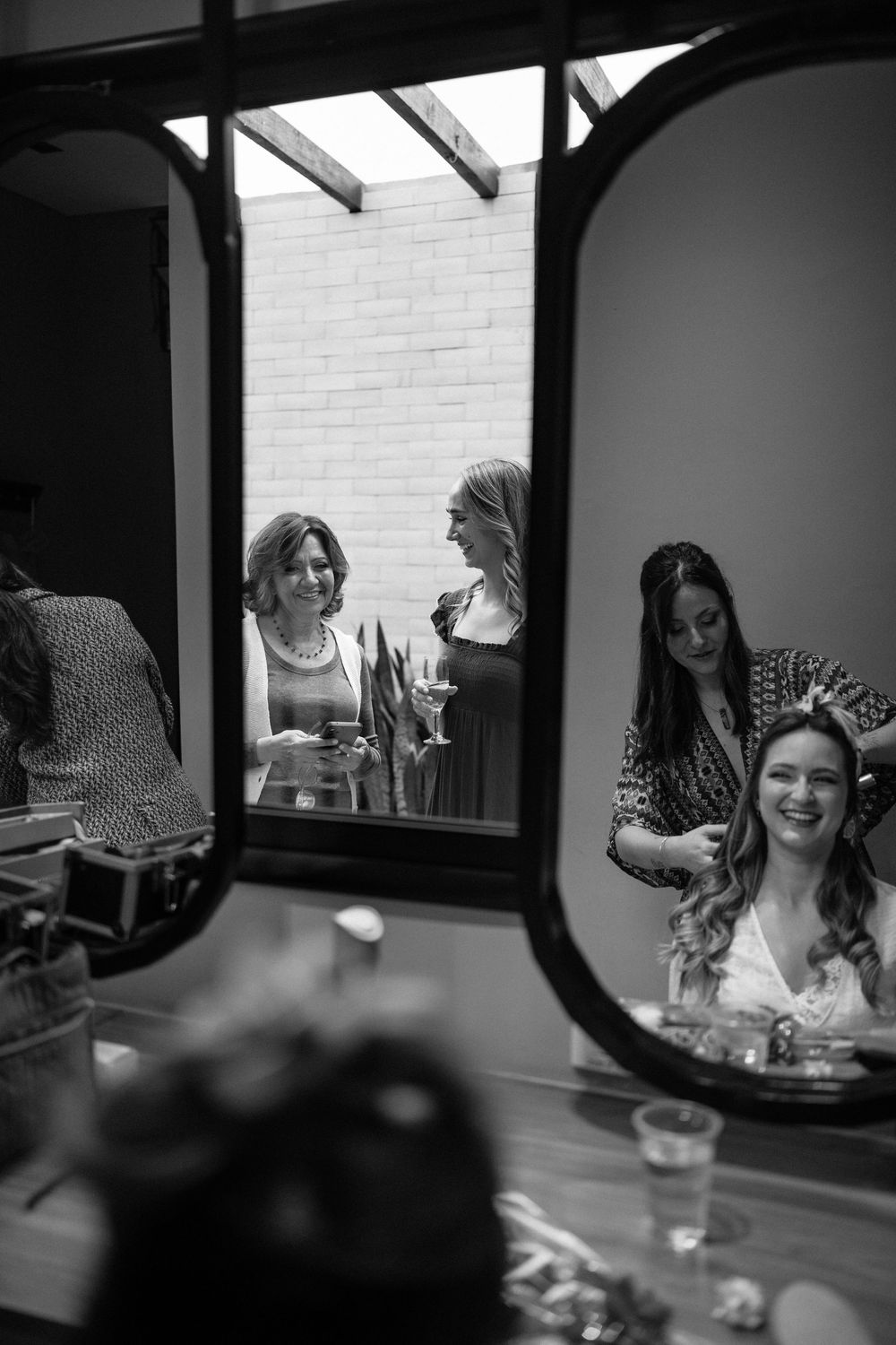 Bride smiling in mirror reflection surrounded by bridesmaids during wedding preparation.