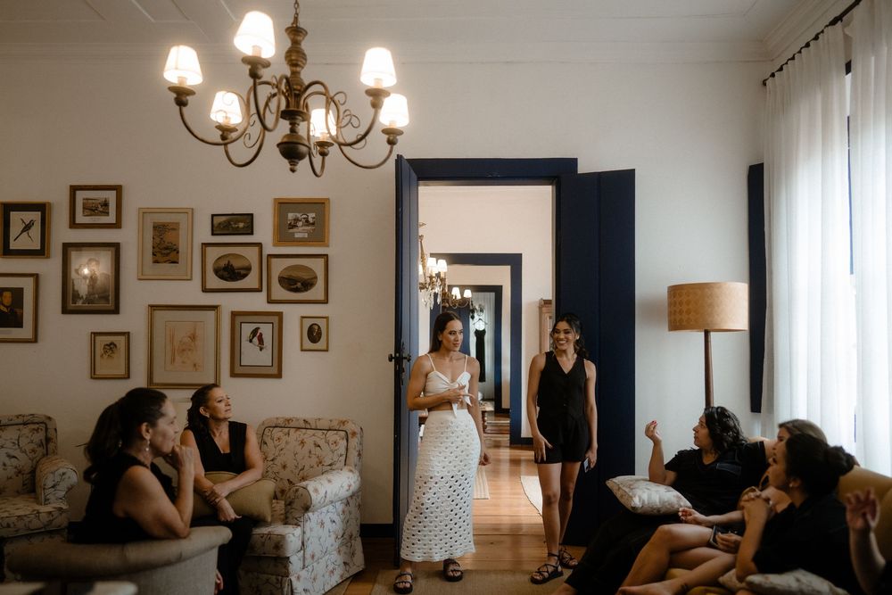 Bride in white dress socializing with bridesmaids in black in elegant room with chandelier.