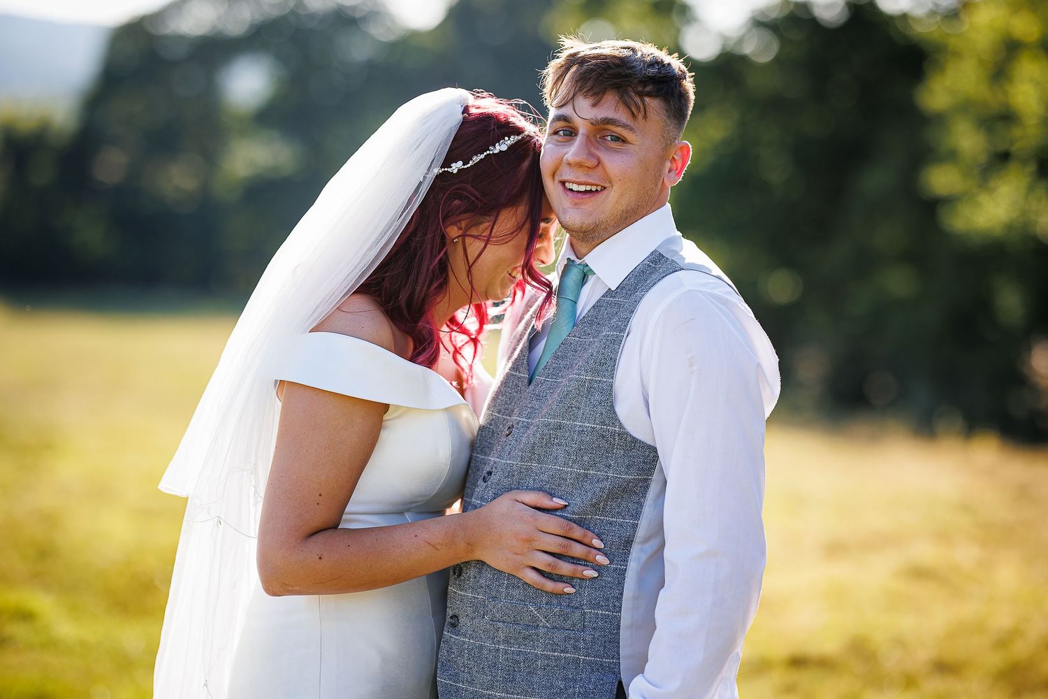 Bride and groom share an intimate moment outdoors, bathed in golden backlight, with lush greenery in the background.