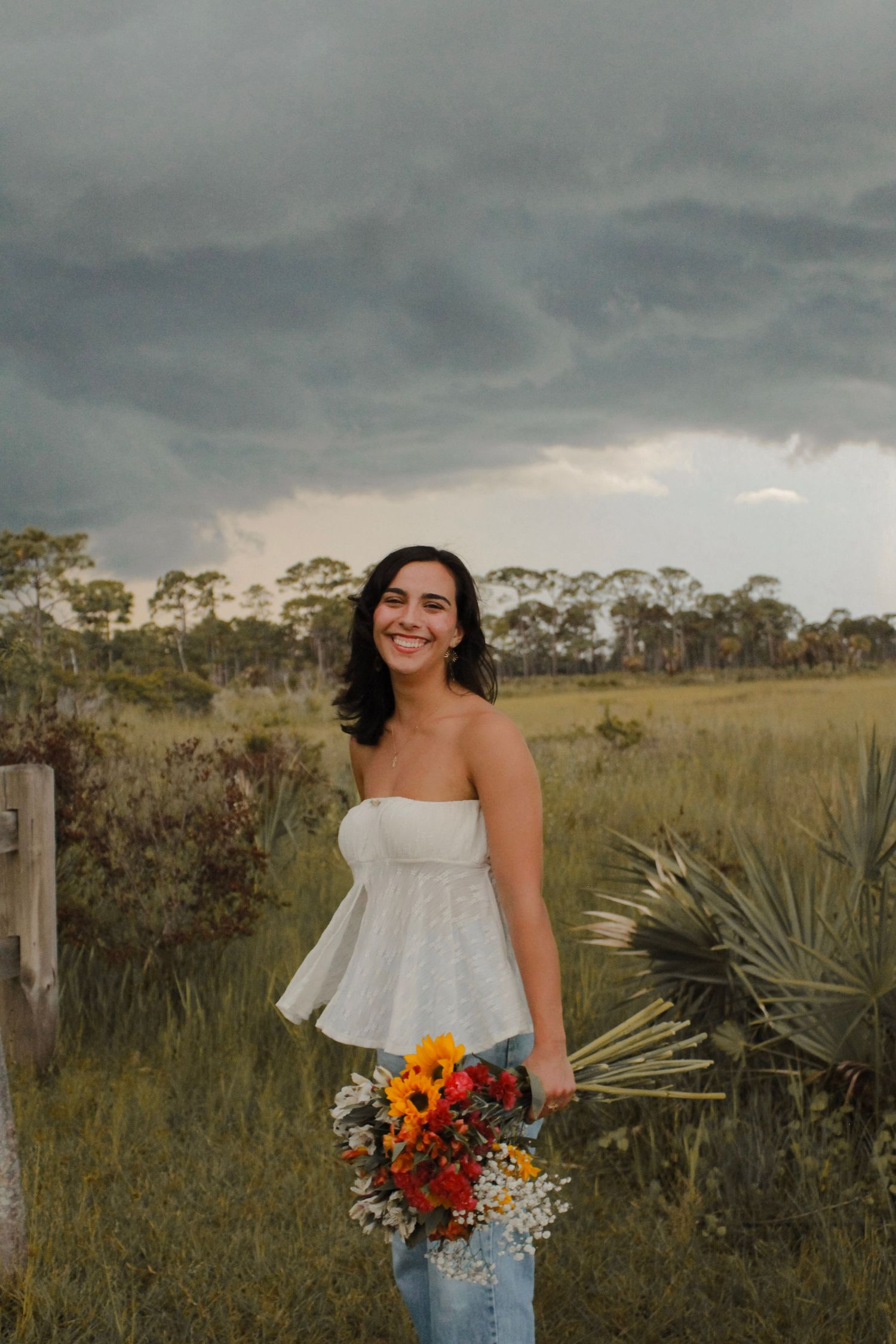 Smiling woman in white strapless top holds colorful bouquet in a field under stormy skies.