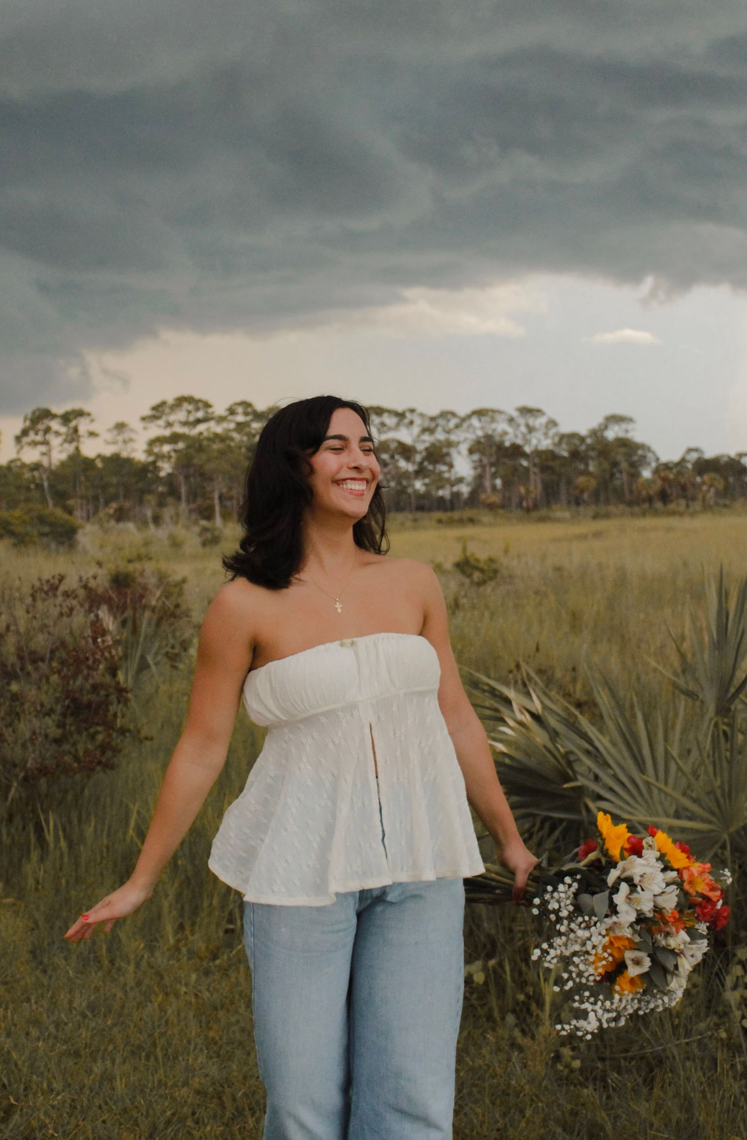 Happy woman in white tube top and jeans holds wildflower bouquet in a grassy field.