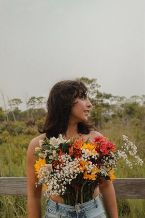 Young woman holds a large colorful bouquet of wildflowers beside a wooden fence outdoors.