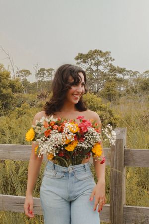 Smiling woman in jeans holds a bright mixed bouquet in front of a rustic wooden fence.