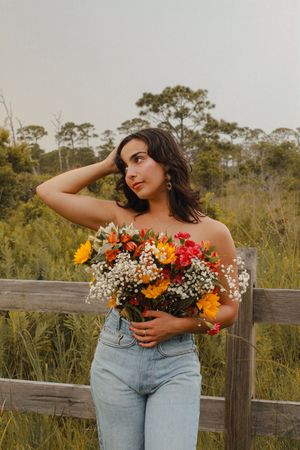 Woman in jeans holds colorful bouquet while posing thoughtfully by a fence in a meadow.