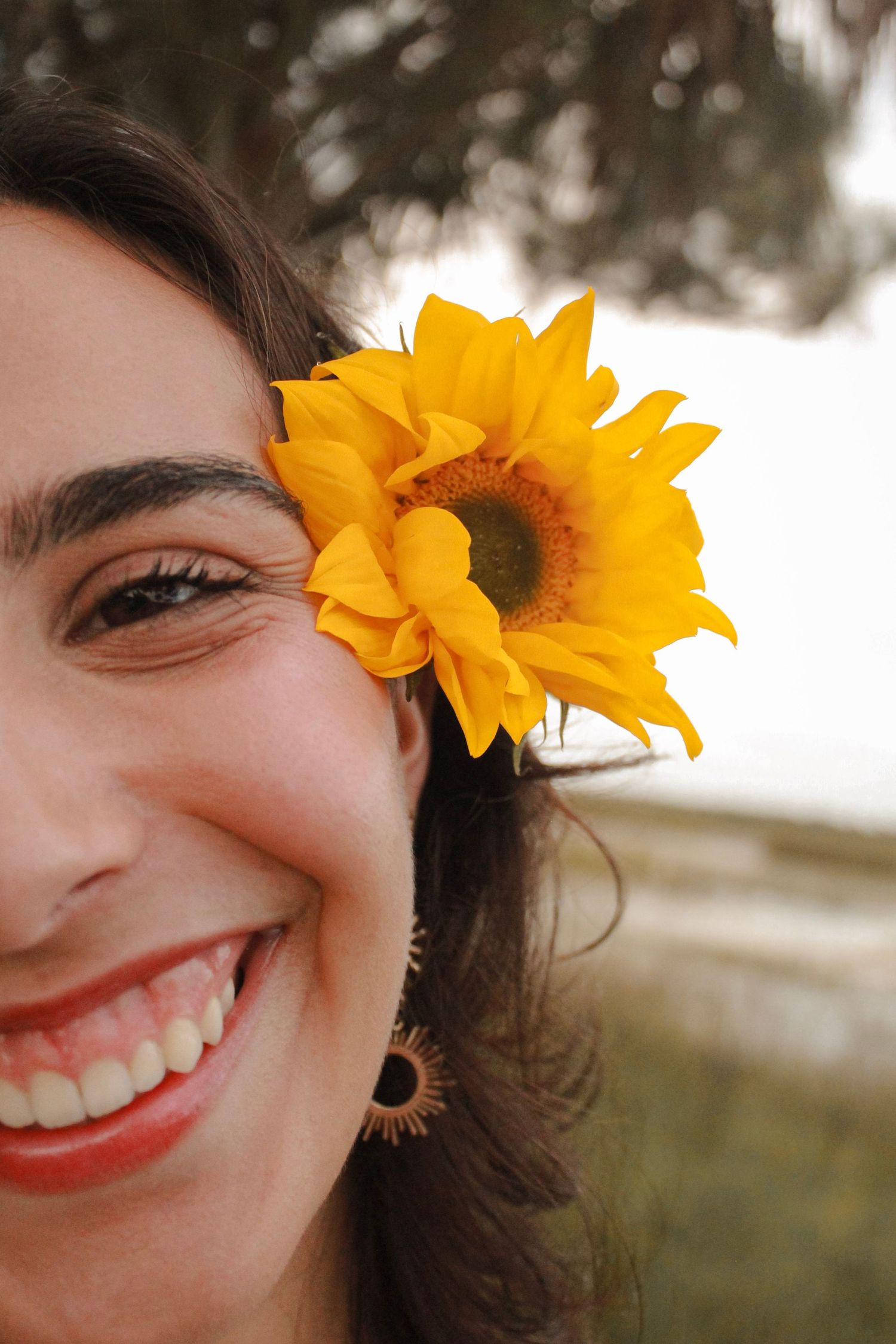 Smiling woman with a bright yellow sunflower tucked in her dark hair, wearing a decorative earring outdoors.