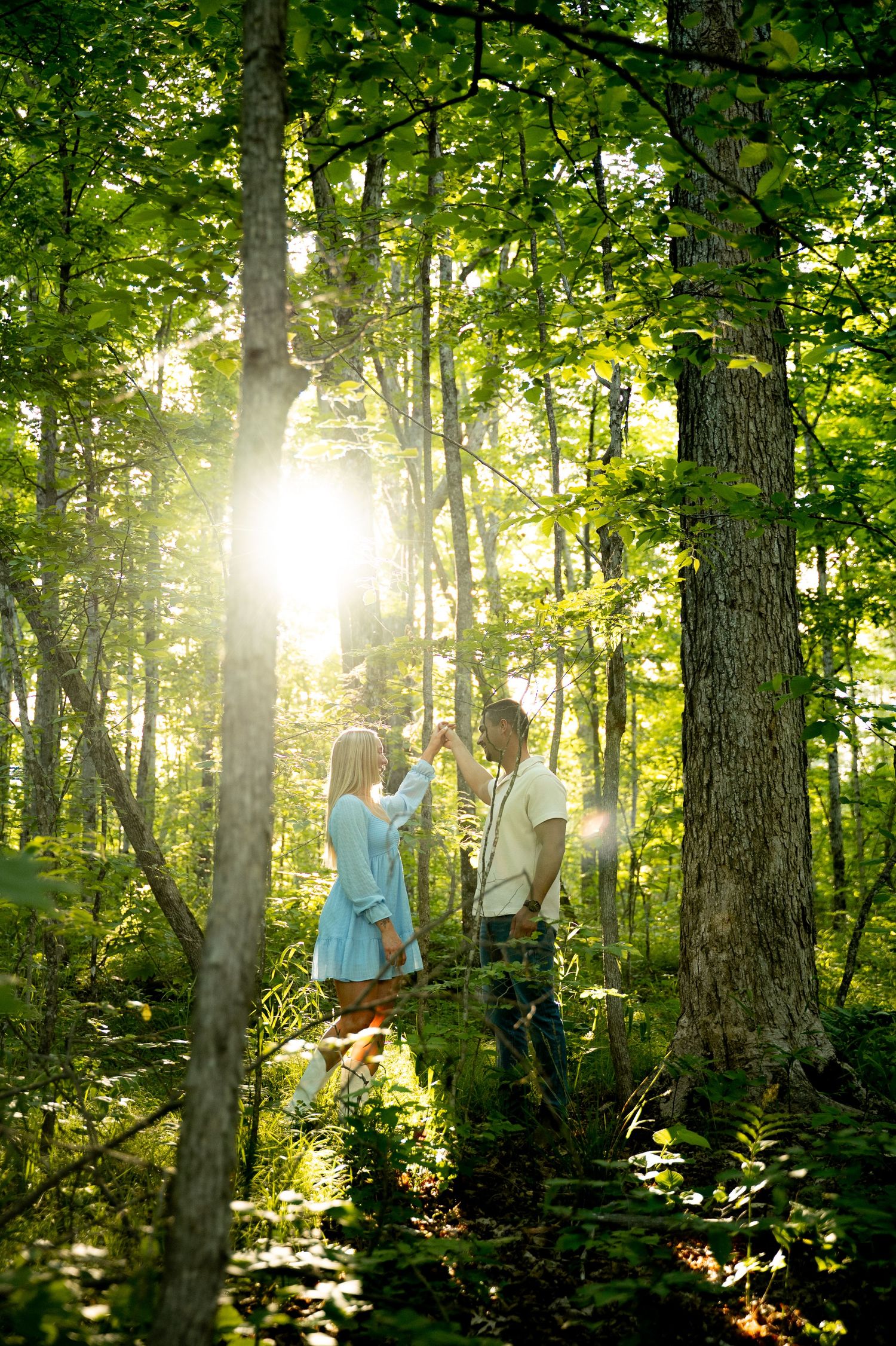 Couple dancing in sunlit forest with green trees and ferns, golden light streaming through lush woodland canopy.