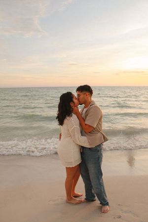 Couple sharing a romantic kiss on a sandy beach at sunset with ocean waves in the background.