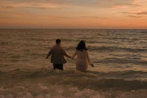 Couple holding hands walking into ocean waves during a vibrant sunset at the beach.
