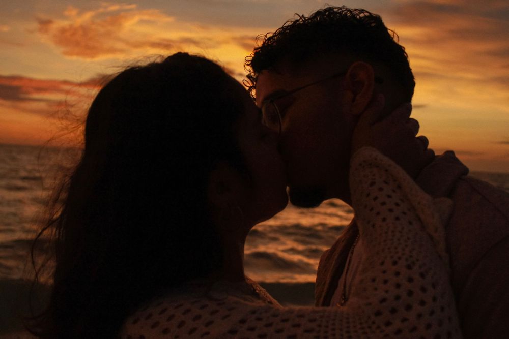 Romantic silhouette of a couple kissing at sunset with glowing orange clouds over the ocean.
