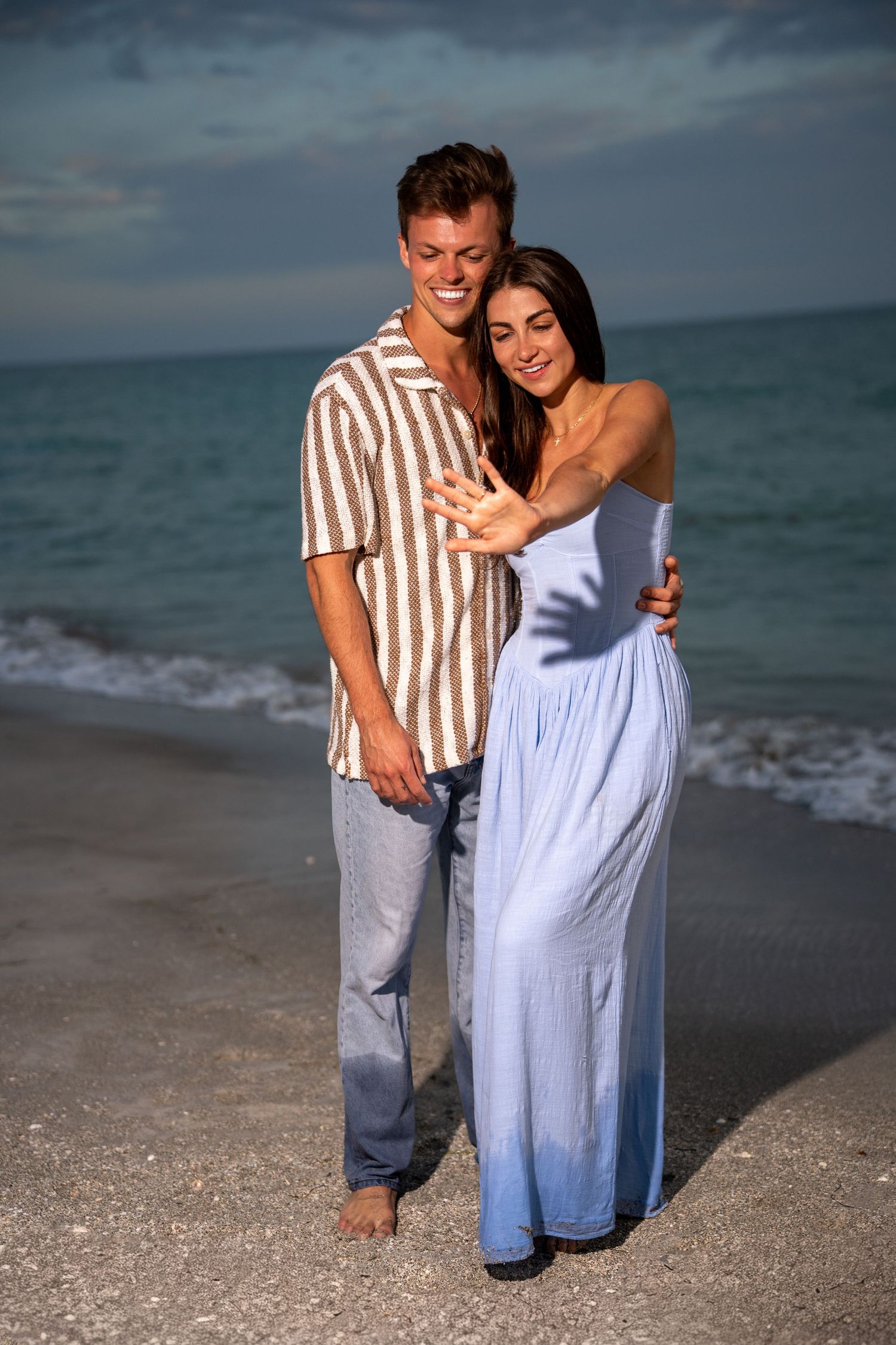 A smiling couple embraces on a sandy beach at sunset, woman in blue dress showing off engagement ring.