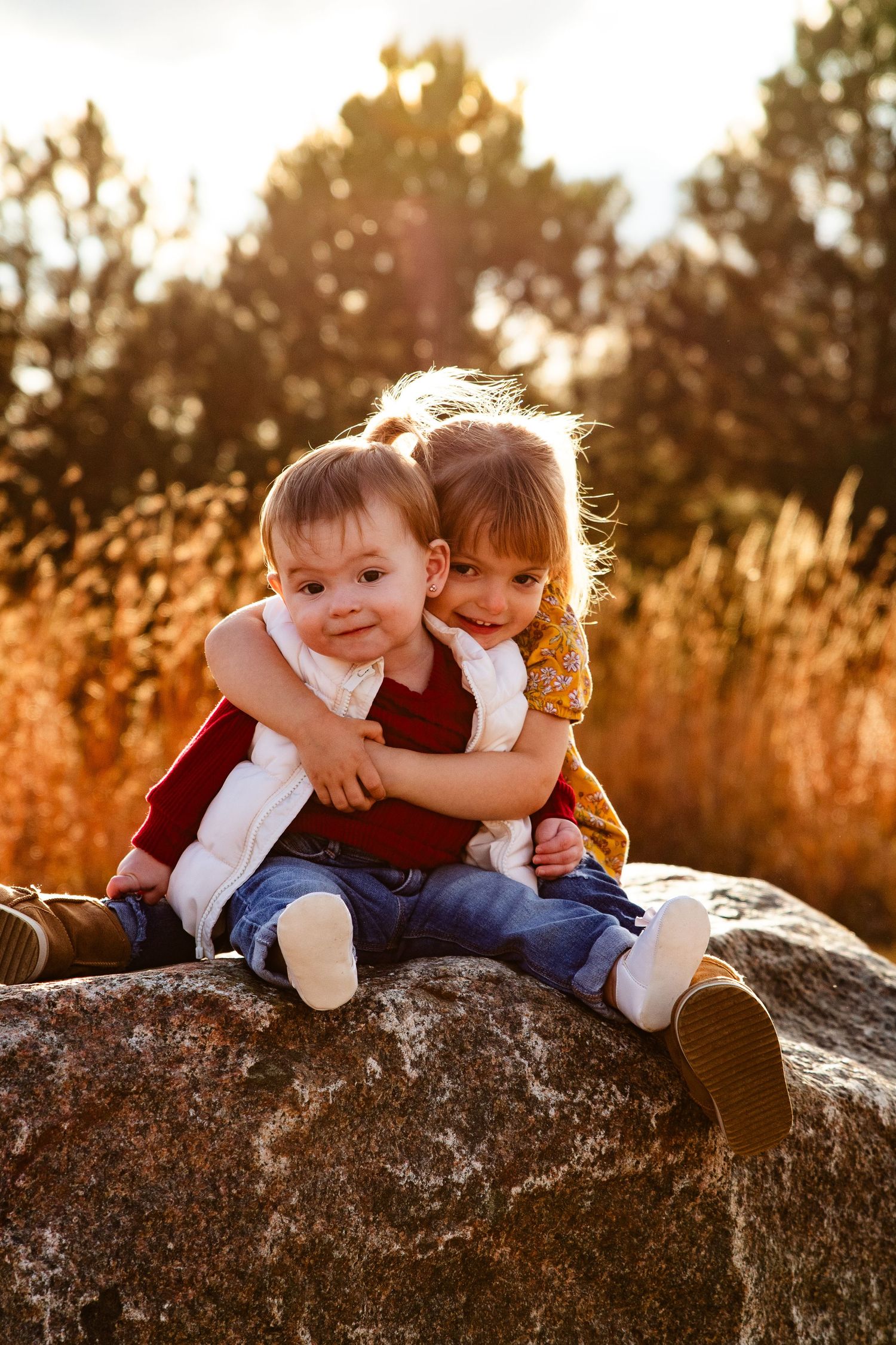 Two young siblings hugging on a rock outdoors during golden hour, surrounded by autumn foliage.