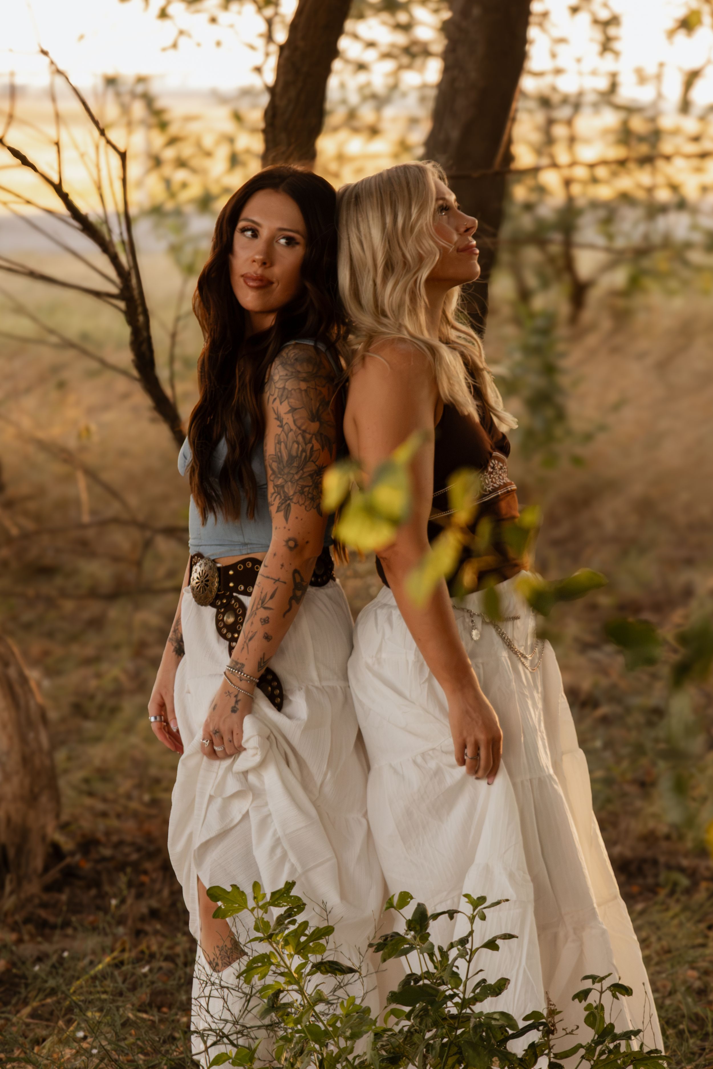 Two women in flowing white dresses posing together outdoors during a creative golden hour portrait session.