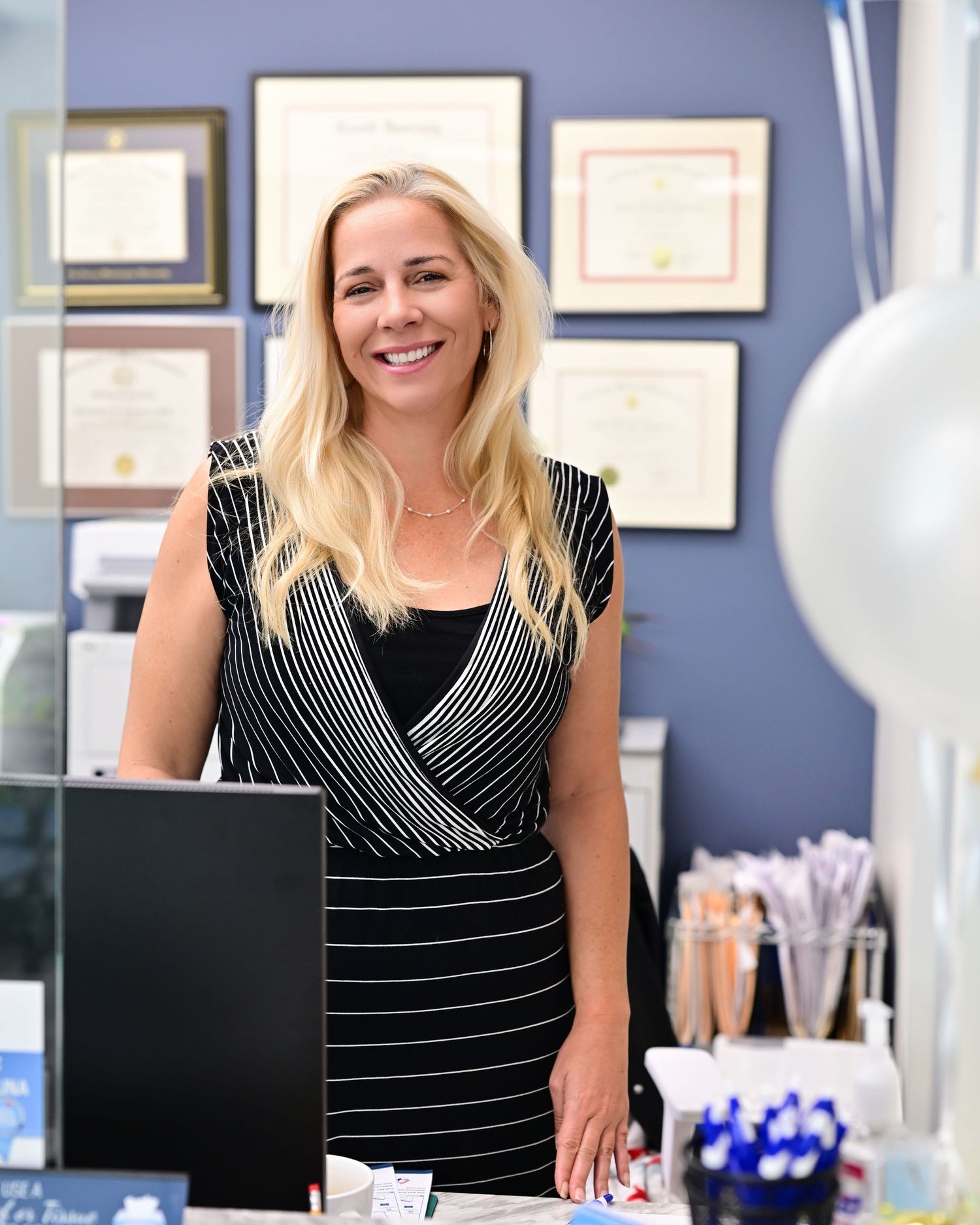Smiling blonde woman in black striped dress standing at reception desk with framed diplomas on wall behind her.