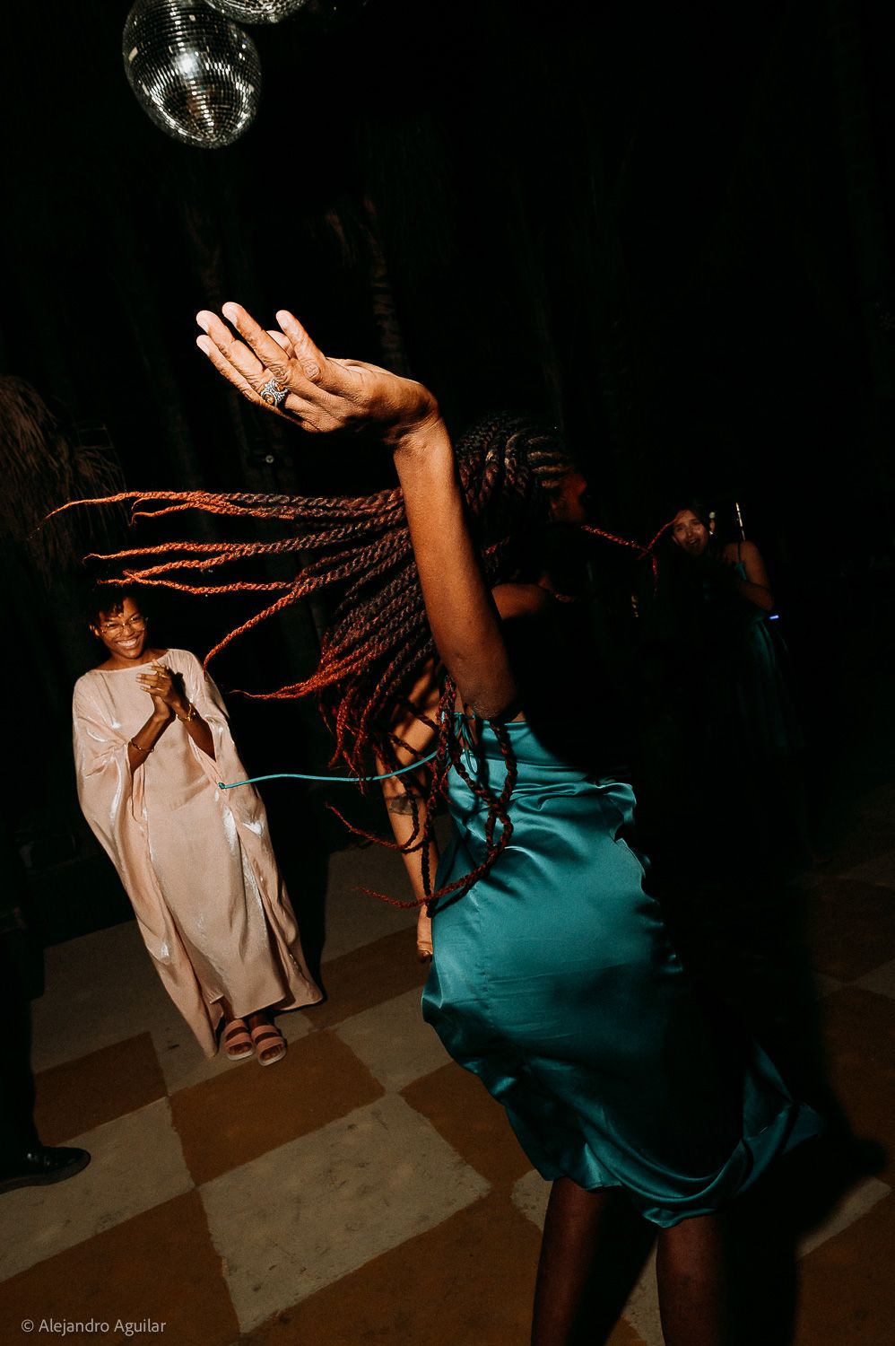 Woman in teal dress dancing energetically with braids flying at a dimly lit party with disco ball overhead.